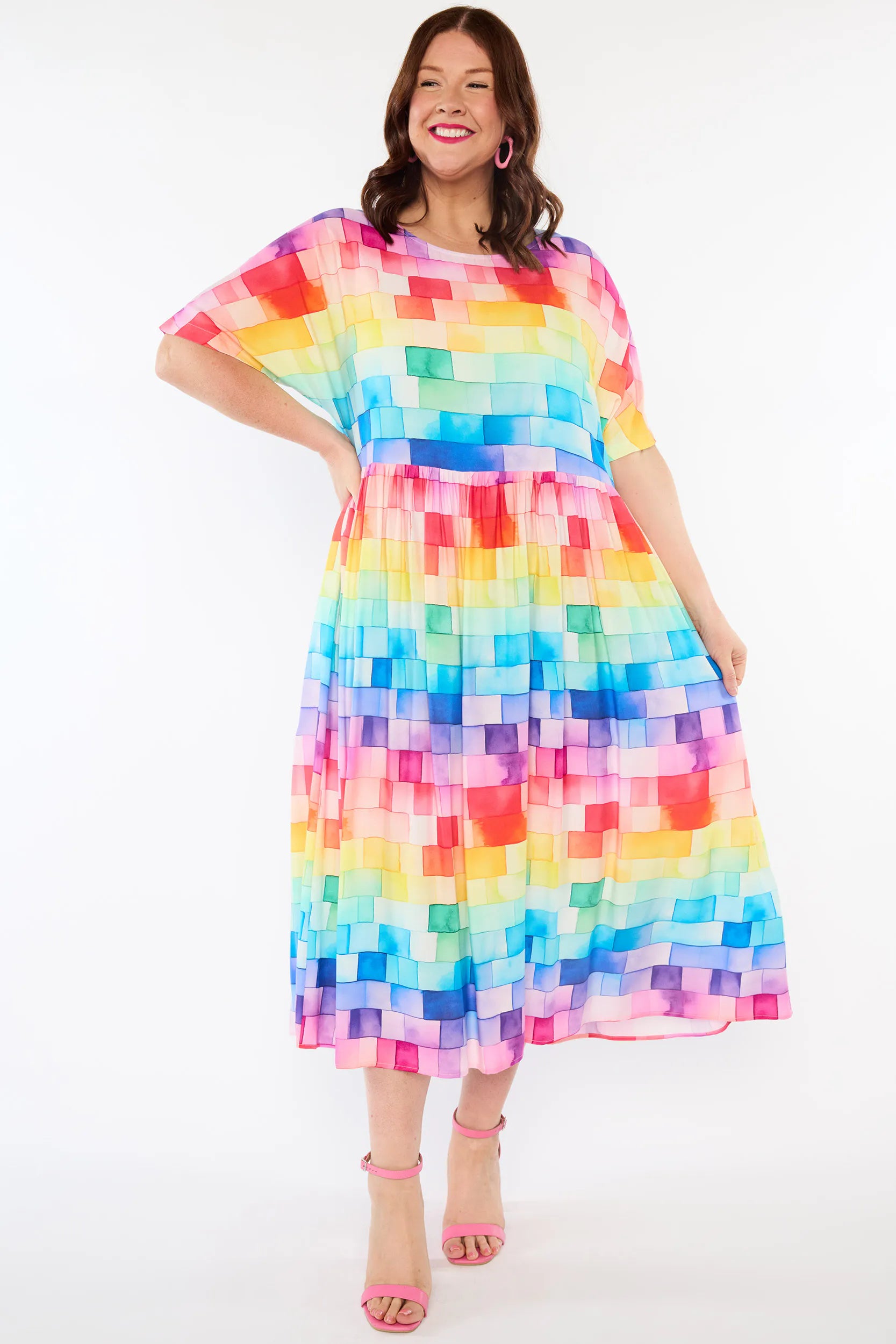 A woman modeling a short-sleeve midi dress with a colorful pixelated block pattern in shades of blue, green, yellow, orange, pink, and purple, paired with pink high-heeled sandals against a white studio background.