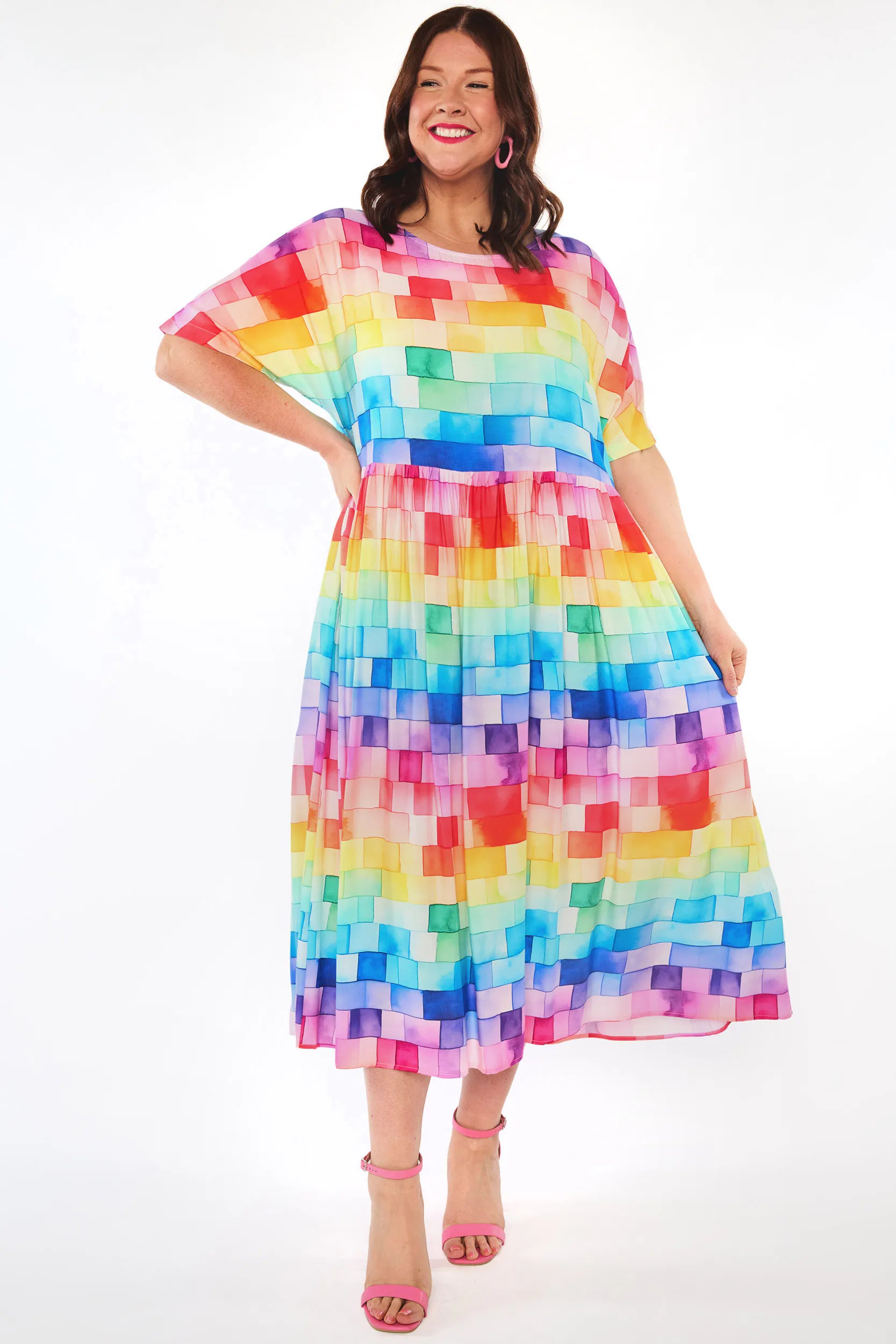 A woman modeling a short-sleeve midi dress with a colorful pixelated block pattern in shades of blue, green, yellow, orange, pink, and purple, paired with pink high-heeled sandals against a white studio background.