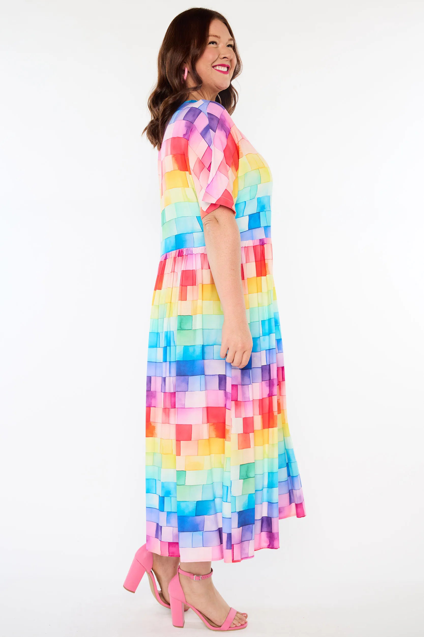 A woman modelling a short-sleeve maxi dress with a colourful geometric pixelated pattern in shades of pink, purple, blue, yellow, and green, paired with pink high-heeled sandals against a white studio background.
