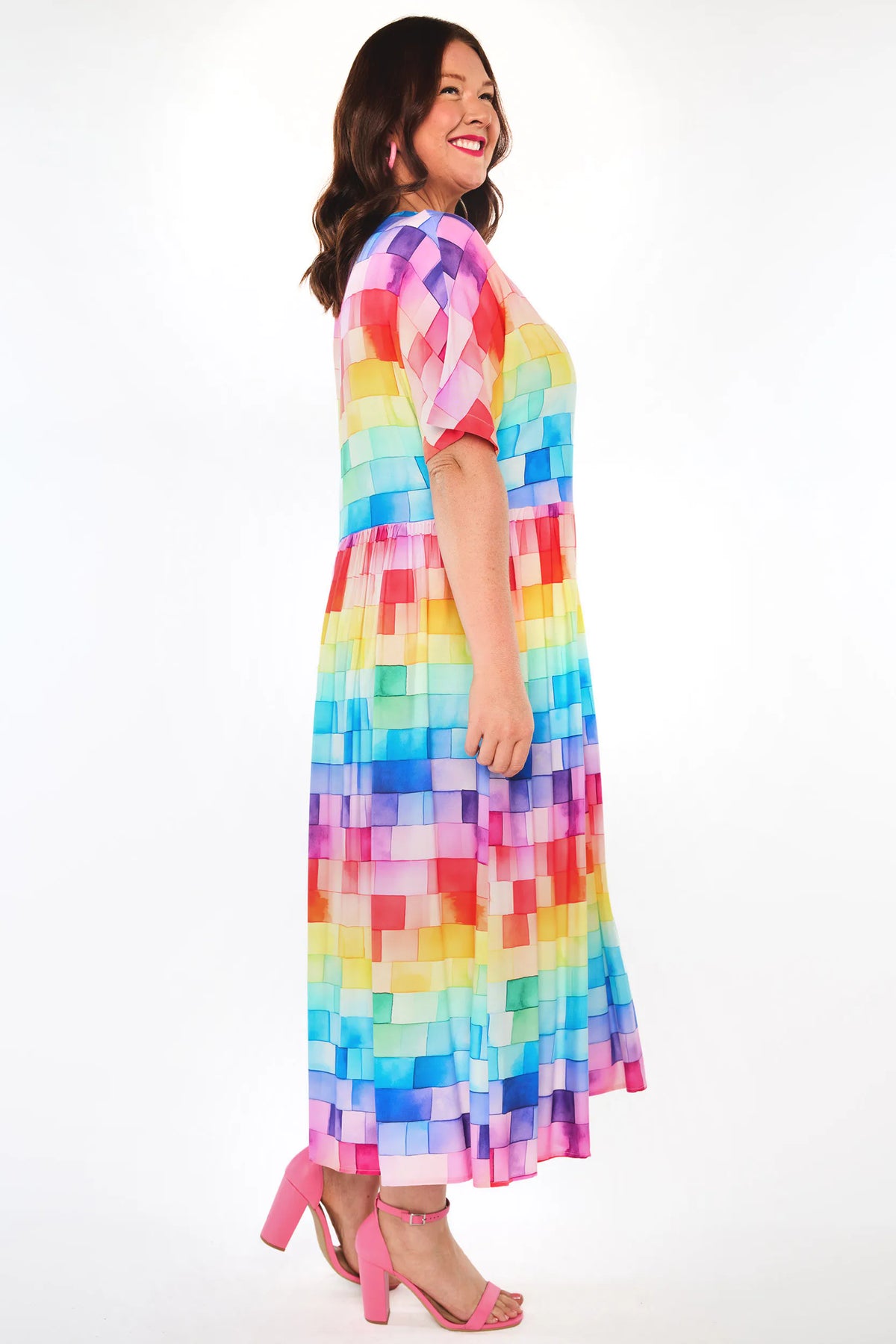 A woman modelling a short-sleeve maxi dress with a colourful geometric pixelated pattern in shades of pink, purple, blue, yellow, and green, paired with pink high heels against a white studio background.