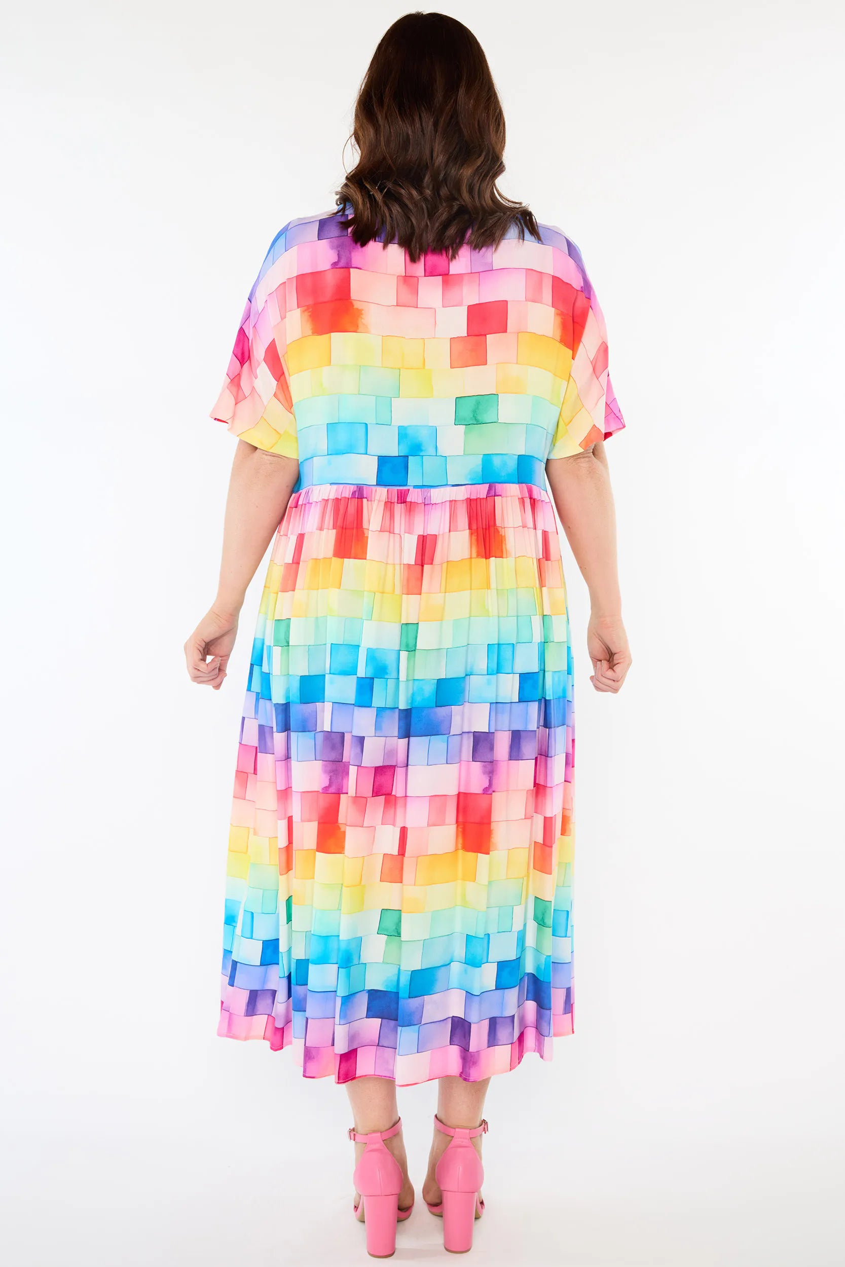 A woman viewed from the back wearing a short-sleeve midi dress with a colourful pixelated block pattern in shades of pink, yellow, orange, green, blue, and purple, paired with pink high heels, against a plain white background.