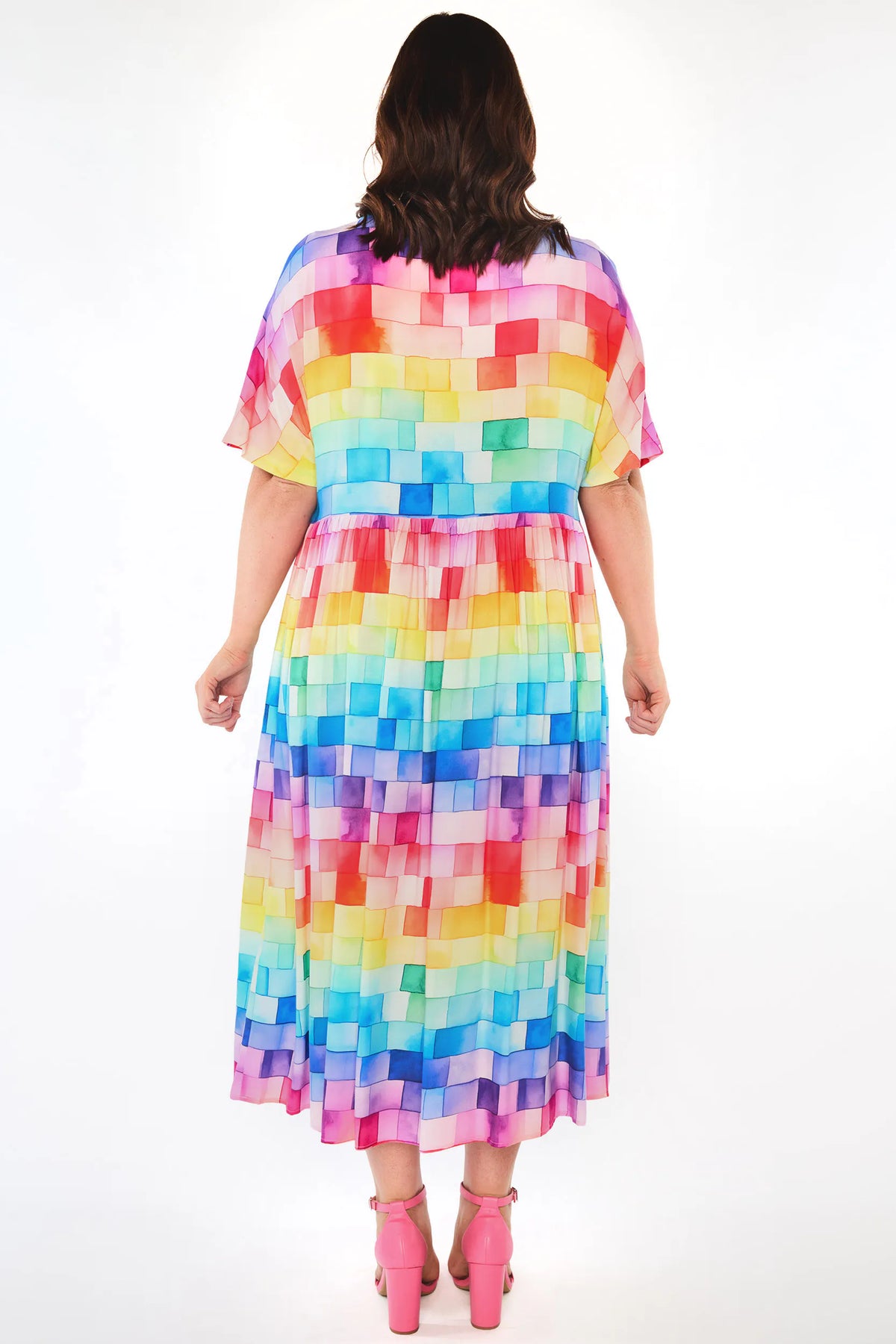 A woman viewed from the back wearing a short-sleeve midi dress with a colorful pixelated block pattern in rainbow shades, paired with pink high heels, against a plain white studio background.