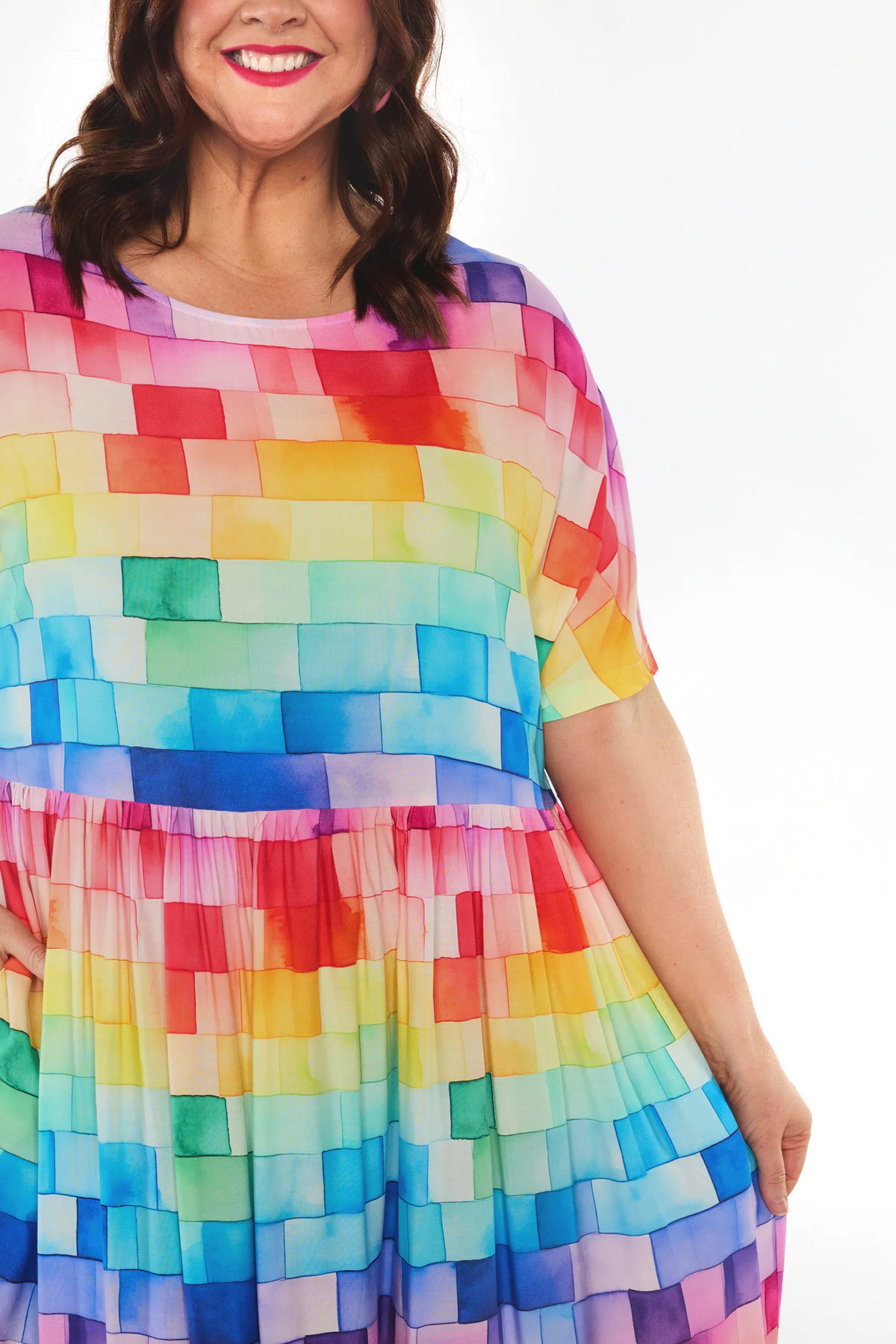 A woman wearing a short-sleeve, tiered dress with a colourful watercolour mosaic pattern of squares in red, orange, yellow, green, blue, and purple, set against a plain white background.