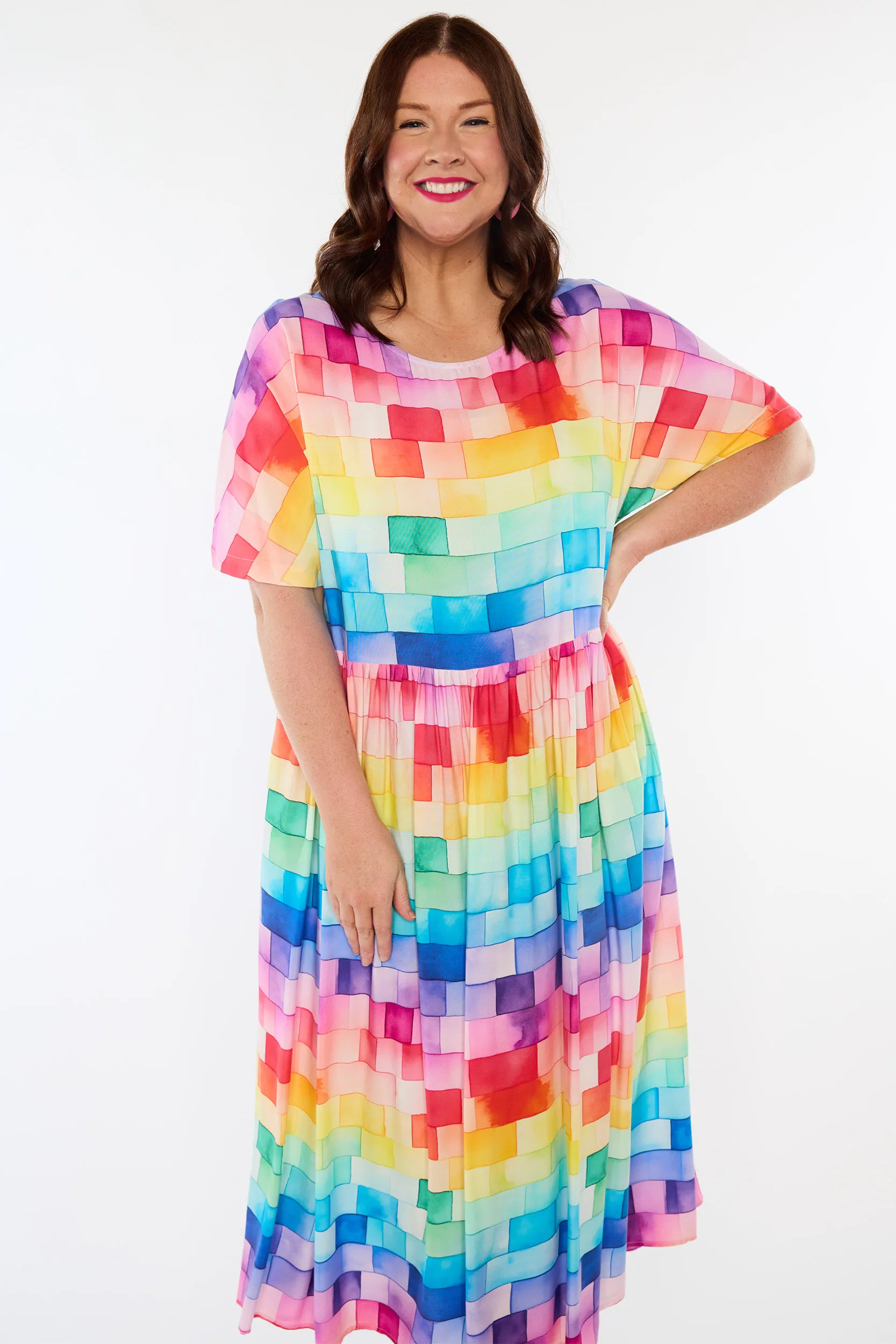 A woman modeling a short-sleeve midi dress with a colorful pixelated block pattern in shades of pink, yellow, orange, green, blue, and purple against a plain white studio background.