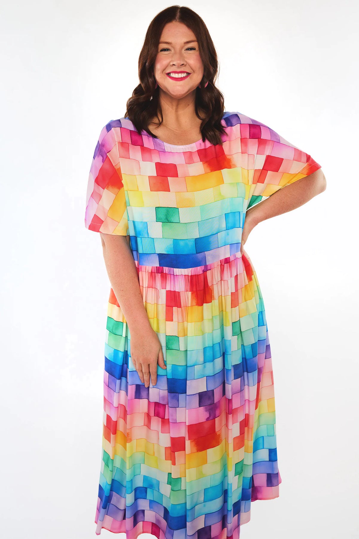 A woman modelling a short-sleeve midi dress with a colourful geometric block pattern in shades of red, orange, yellow, green, blue, and purple, set against a plain white studio background.