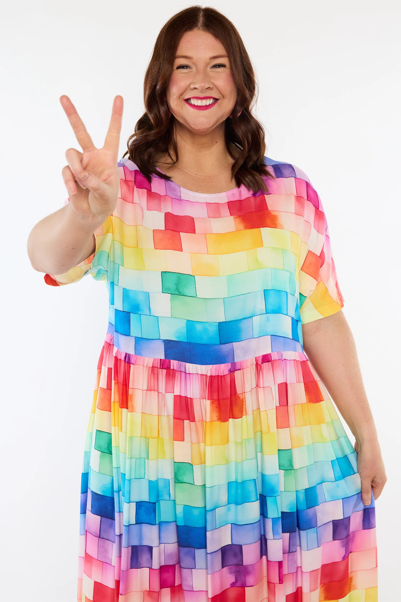 A woman wearing a short-sleeve A-line dress with a colorful rainbow block pattern, posing and making a peace sign against a plain white background.