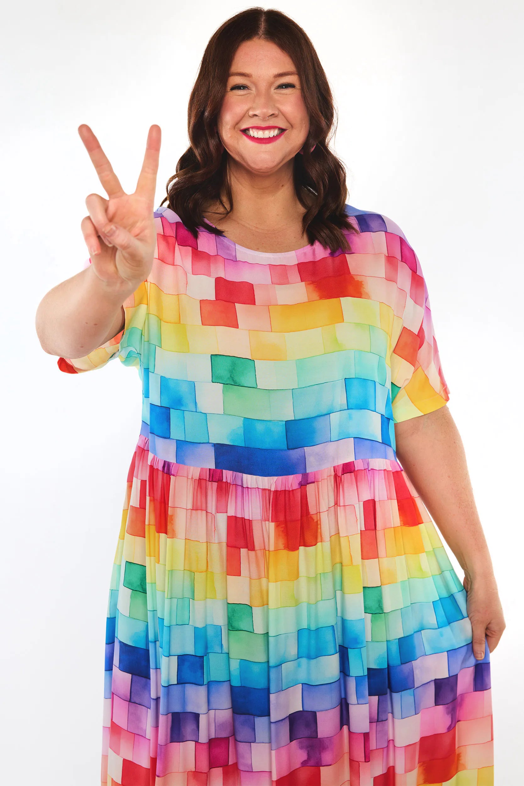 A woman smiling and making a peace sign, wearing a short-sleeve A-line dress with a colourful watercolour block pattern in shades of red, orange, yellow, green, blue, and purple on a white background.