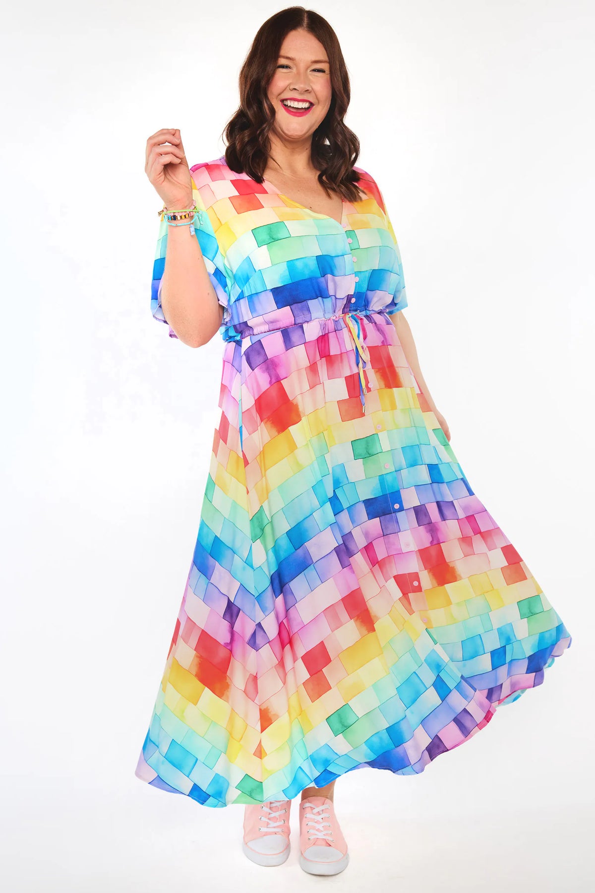 A woman modeling a vibrant, multicolor midi dress with a pixelated block pattern in shades of blue, green, yellow, pink, orange, and purple. She is smiling and posing against a clean white studio background.