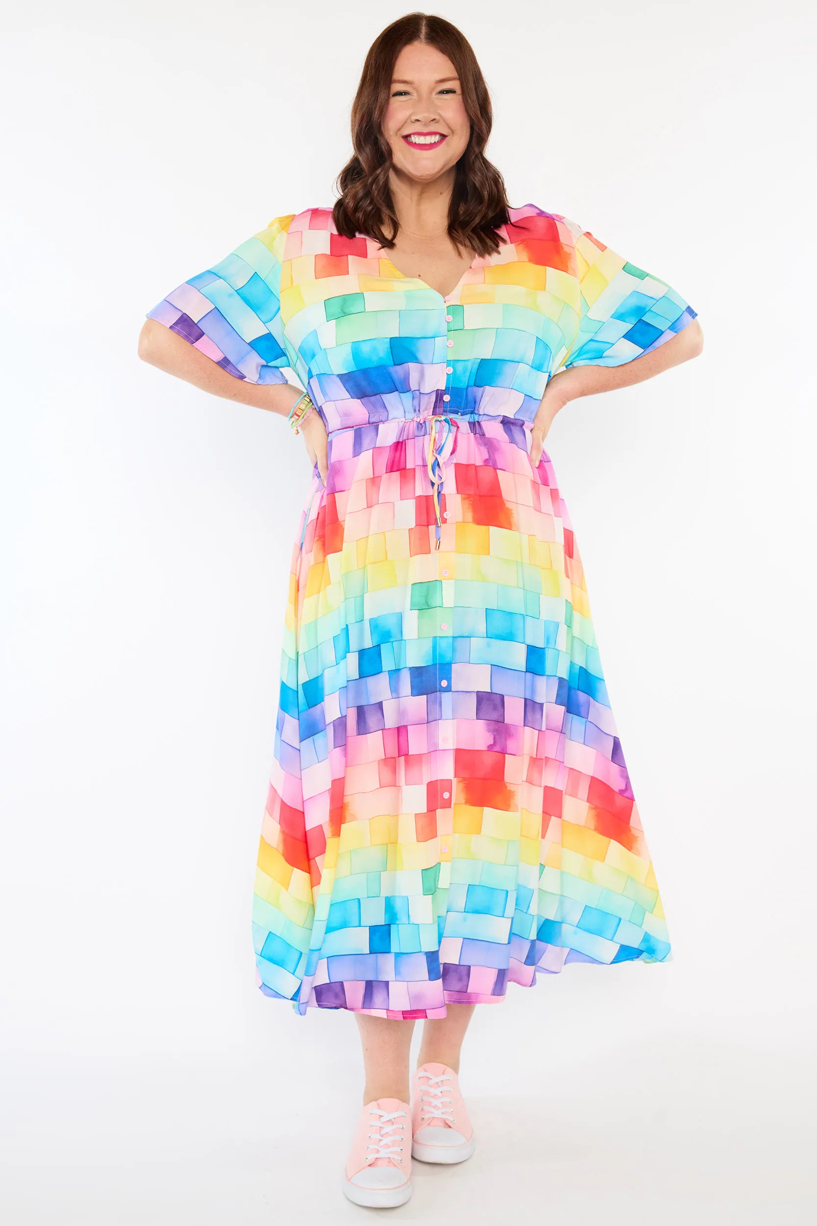 A woman modelling a short-sleeve, Midi-dress dress, with a button-up V-neckline & adjustable tie waist. A rainbow geometric print covered with rainbow squares, paired with pink sandshoes, against a plain white studio background.