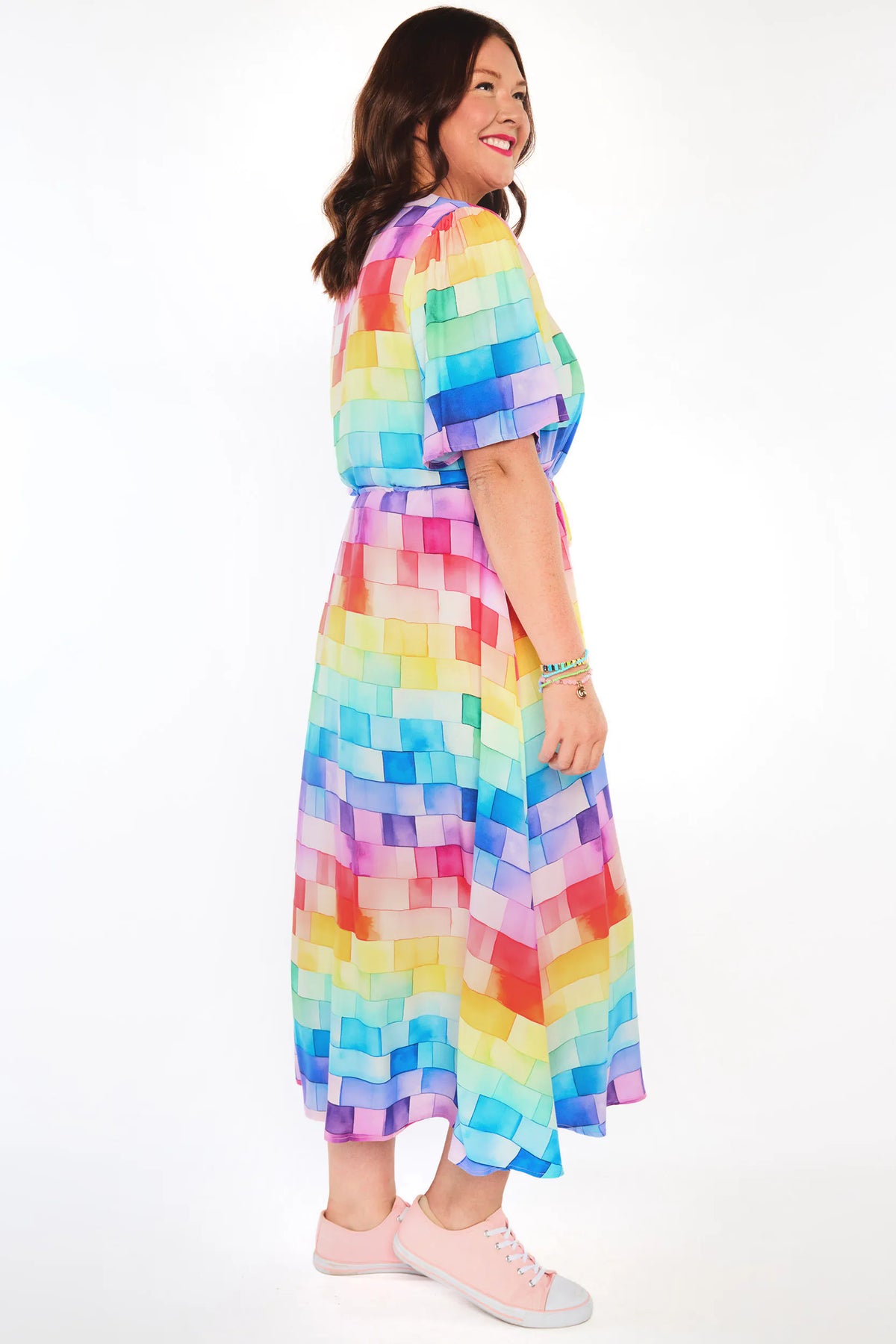A woman modeling a short-sleeve midi dress with a colorful geometric block pattern in shades of blue, yellow, pink, orange, and purple, paired with light pink sneakers against a white studio background.