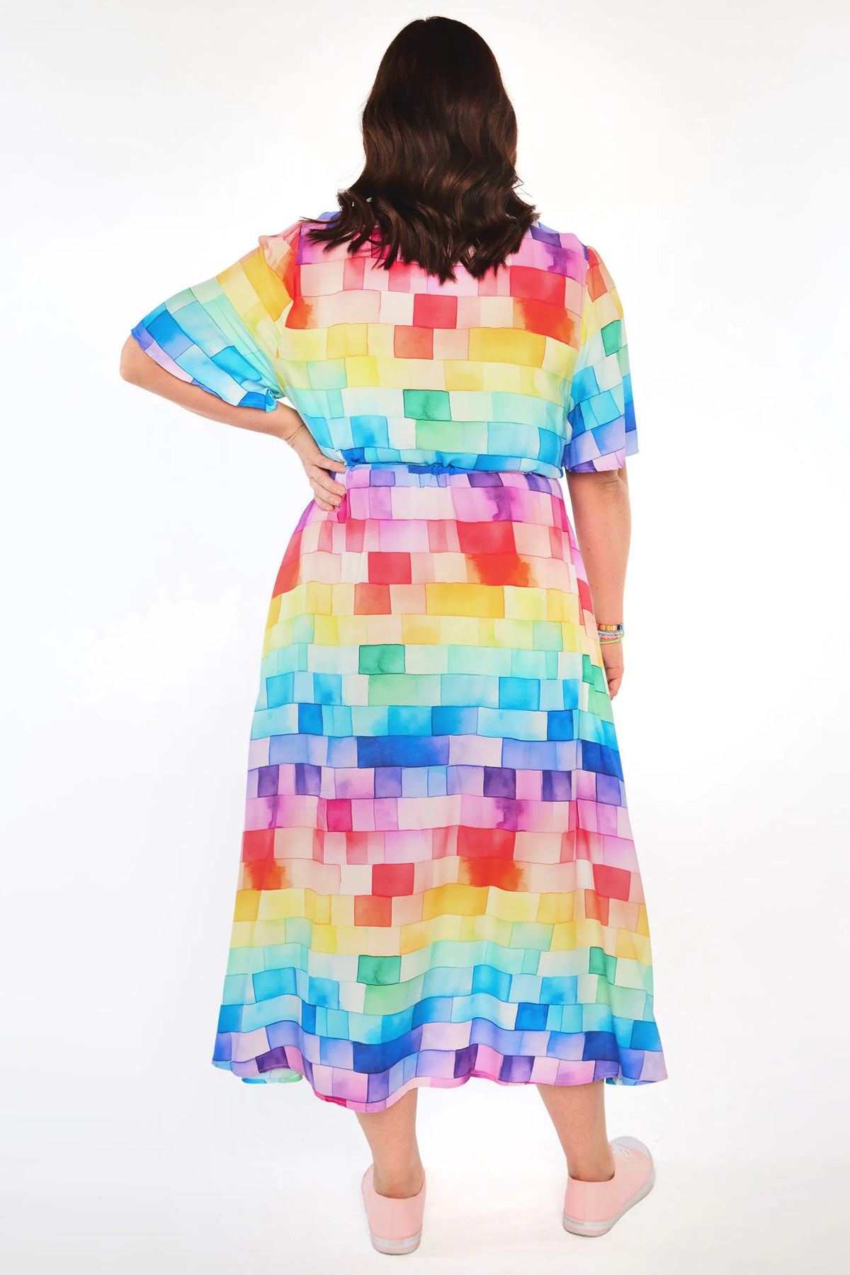 A woman viewed from behind wearing a short-sleeved midi dress with a colorful pixelated block pattern in shades of blue, green, yellow, orange, pink, purple, and red. She is standing against a plain white studio background.