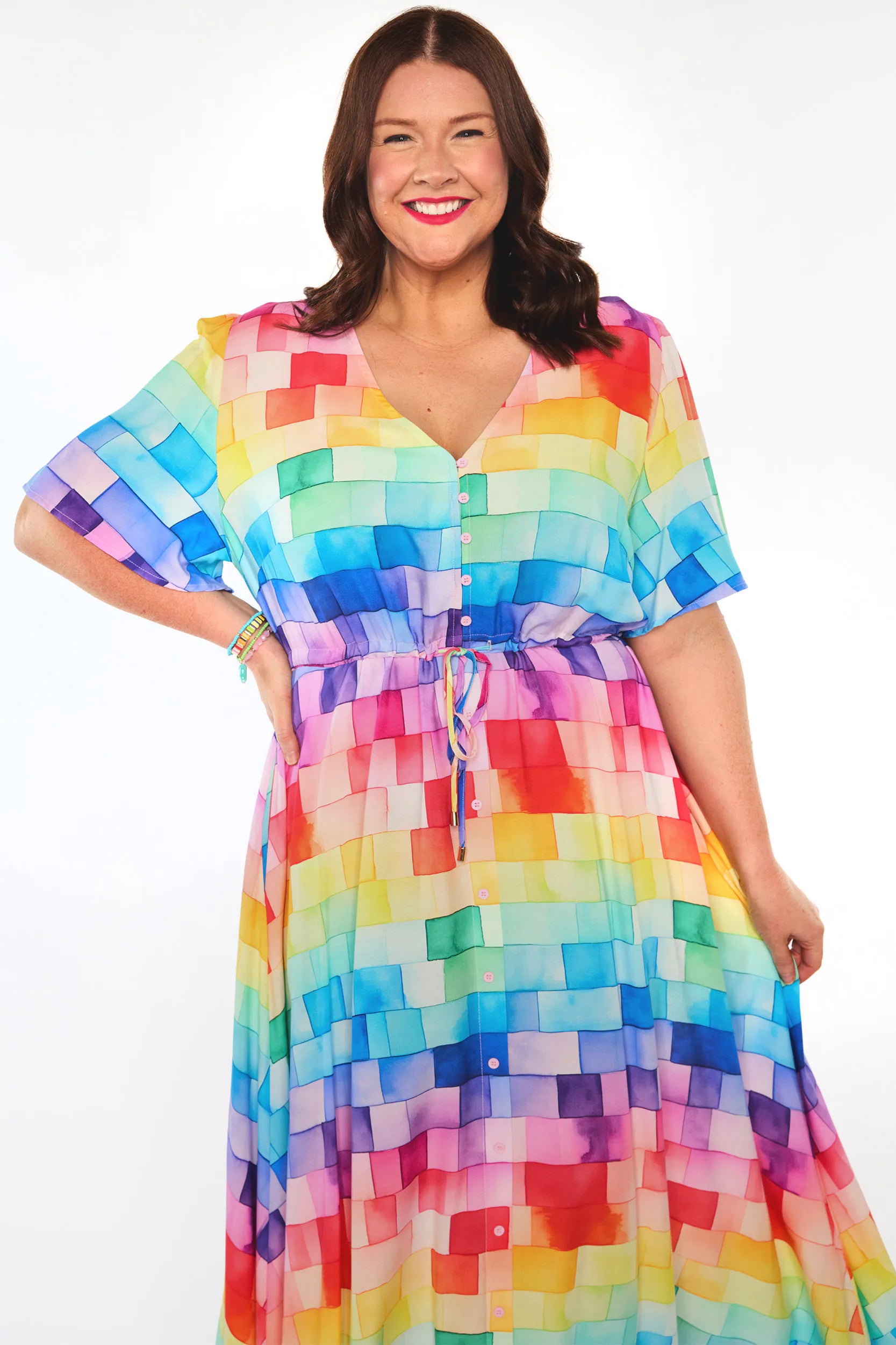 A woman modeling a short-sleeve V-neck dress with a colorful pixelated block pattern in rainbow colors, set against a plain white studio background.