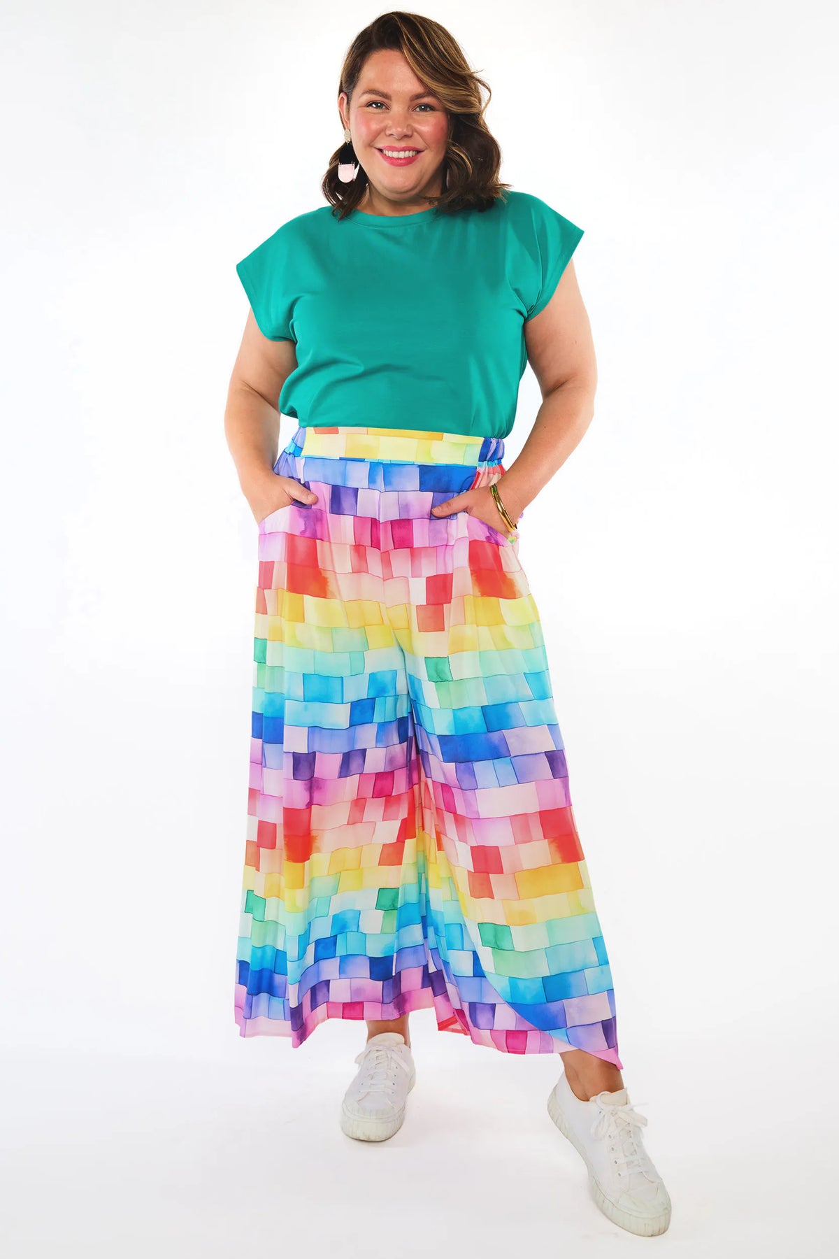 A woman modeling a vibrant, two-piece outfit consisting of a solid teal green short-sleeve top and high-waisted, wide-leg pants with a colorful rainbow pixelated block pattern, paired with white sneakers on a white studio background.