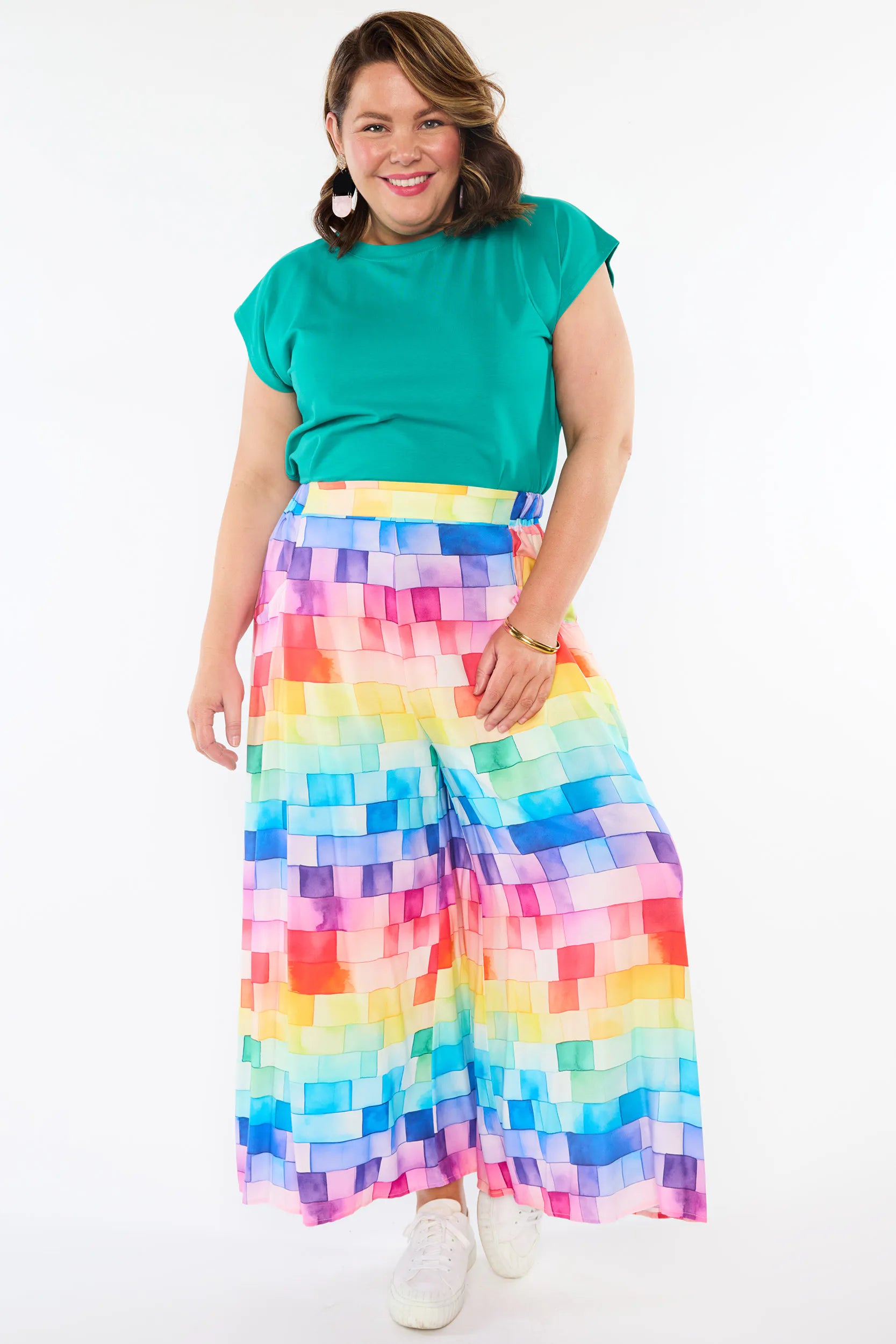 A woman modeling a bright green short-sleeve top and high-waisted, wide-leg pleated pants featuring a colorful rainbow pixelated block pattern, paired with white sneakers against a plain white background.