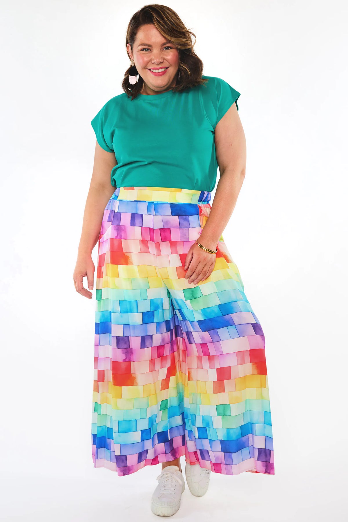 A woman modeling a vibrant, multicolor pixelated print wide-leg skirt paired with a green short-sleeve top and white sneakers, set against a plain white studio background.