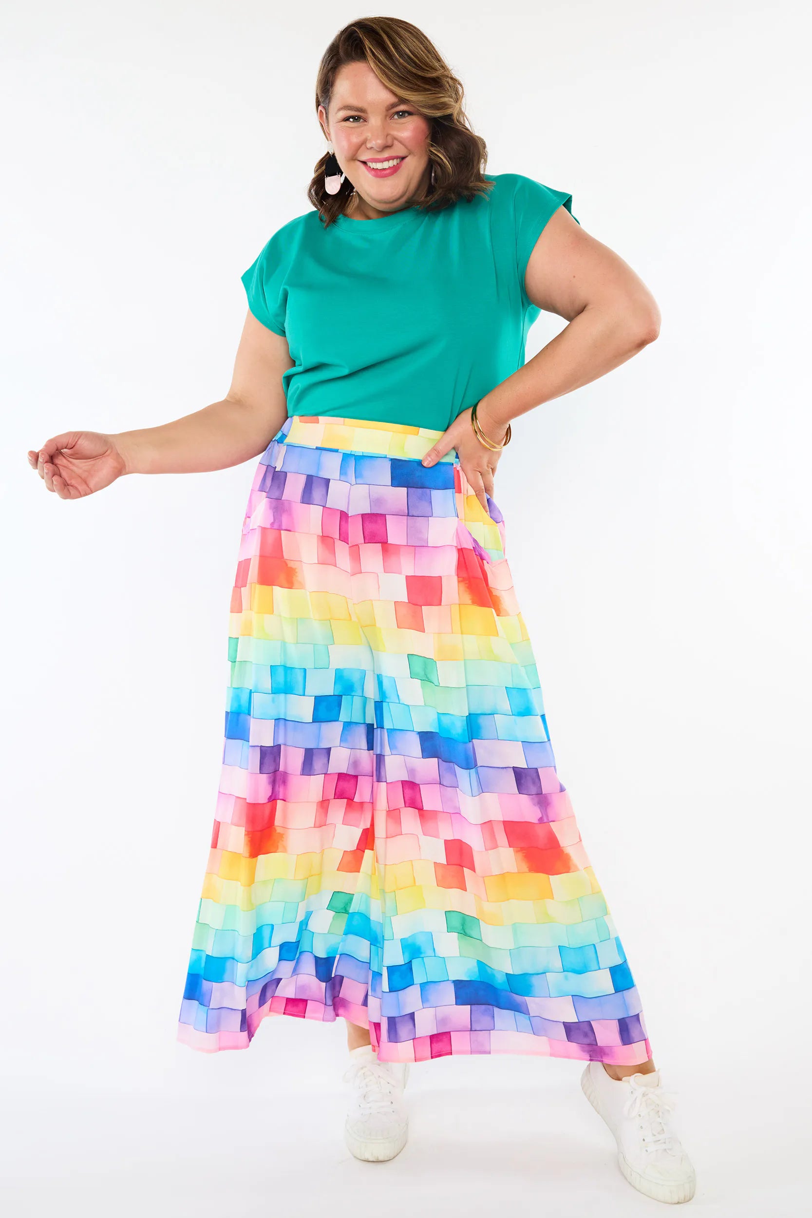 A woman modeling a vibrant, multicolor maxi skirt with a rainbow checkerboard pattern and a solid teal green short-sleeve top, standing against a plain white studio background.