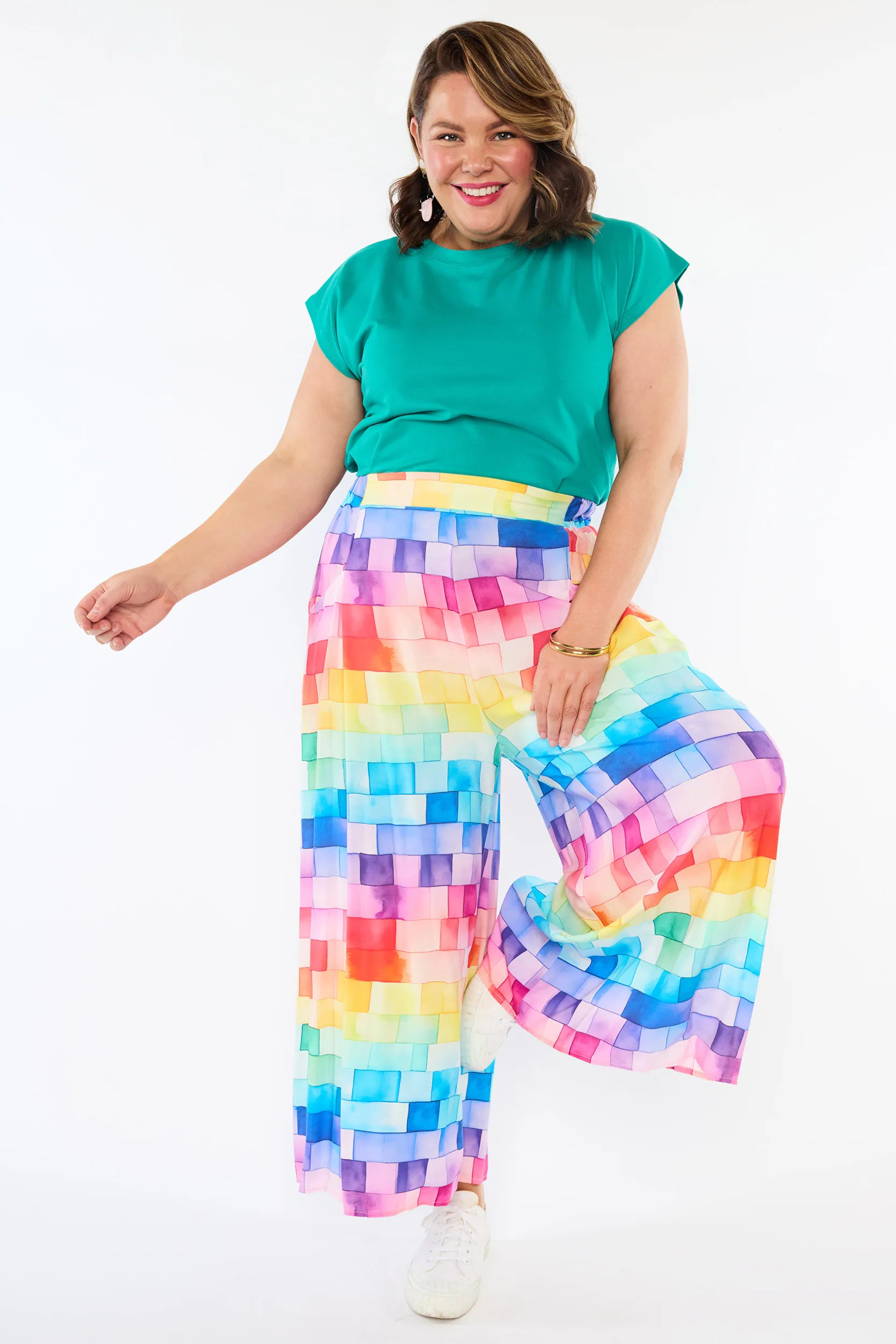 A woman modeling a vibrant, multicolor wide-leg pant with a pixelated block pattern, paired with a solid teal green top and white sneakers, against a plain white studio background.