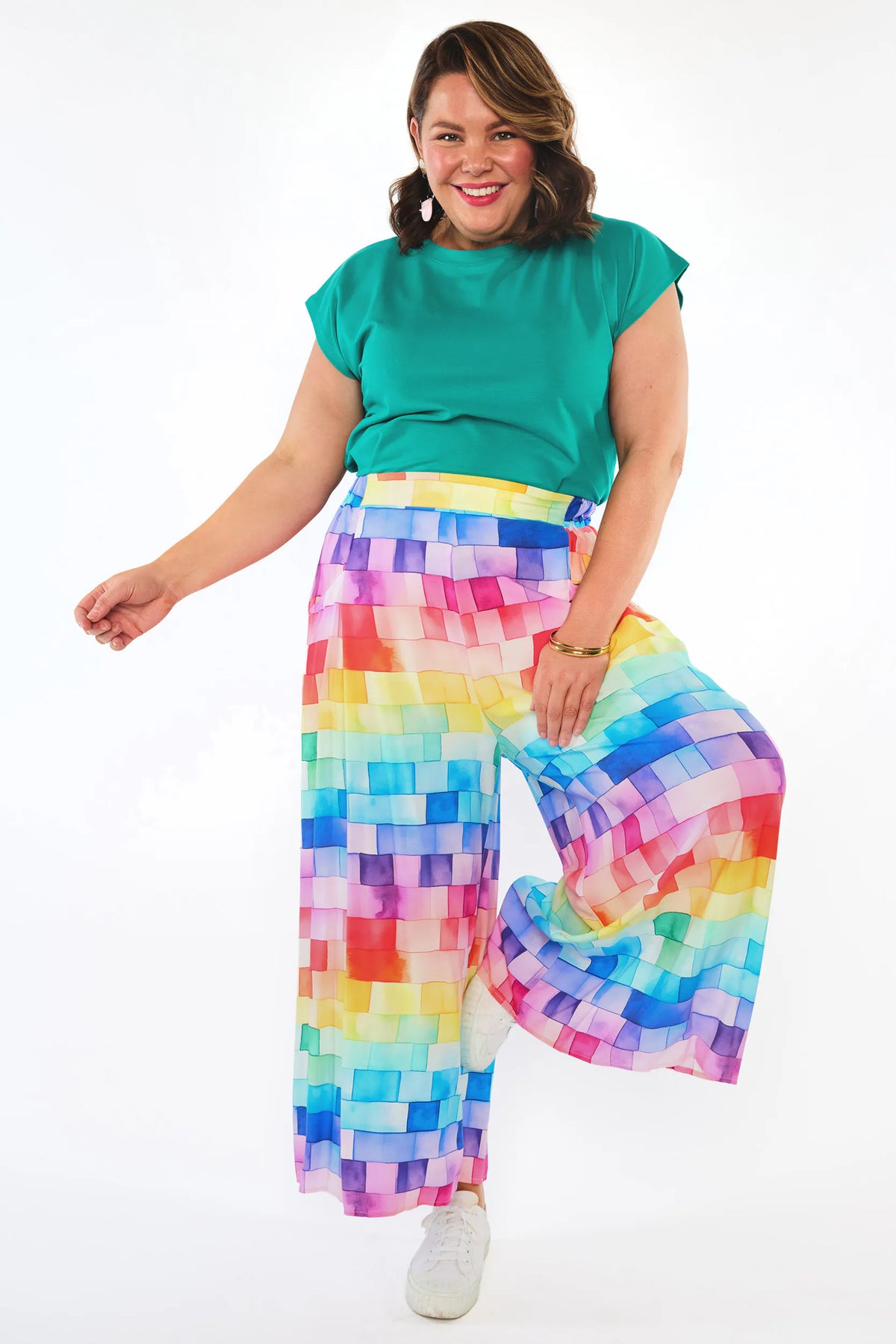 A woman modeling a pair of wide-leg pants with a colorful pixelated pattern, paired with a solid teal green t-shirt and white sneakers, against a plain white background.