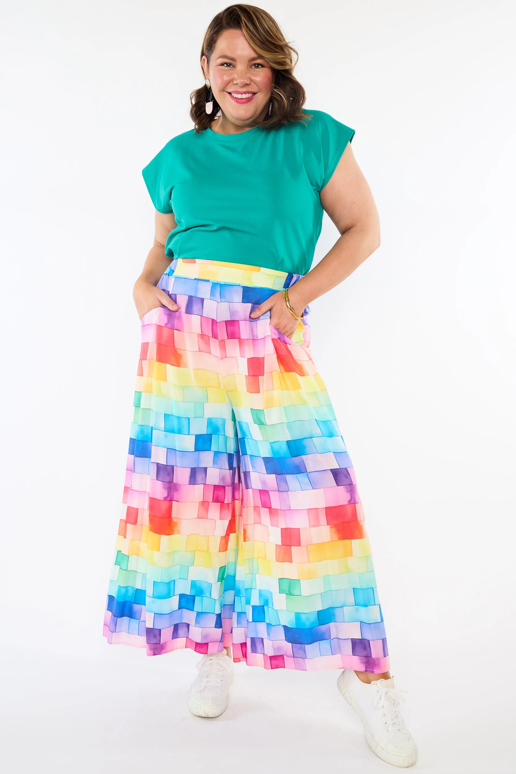 A woman modeling a bright green short-sleeve top and a wide-leg pleated skirt with a colorful pixelated rainbow pattern, paired with white sneakers, against a plain white background.