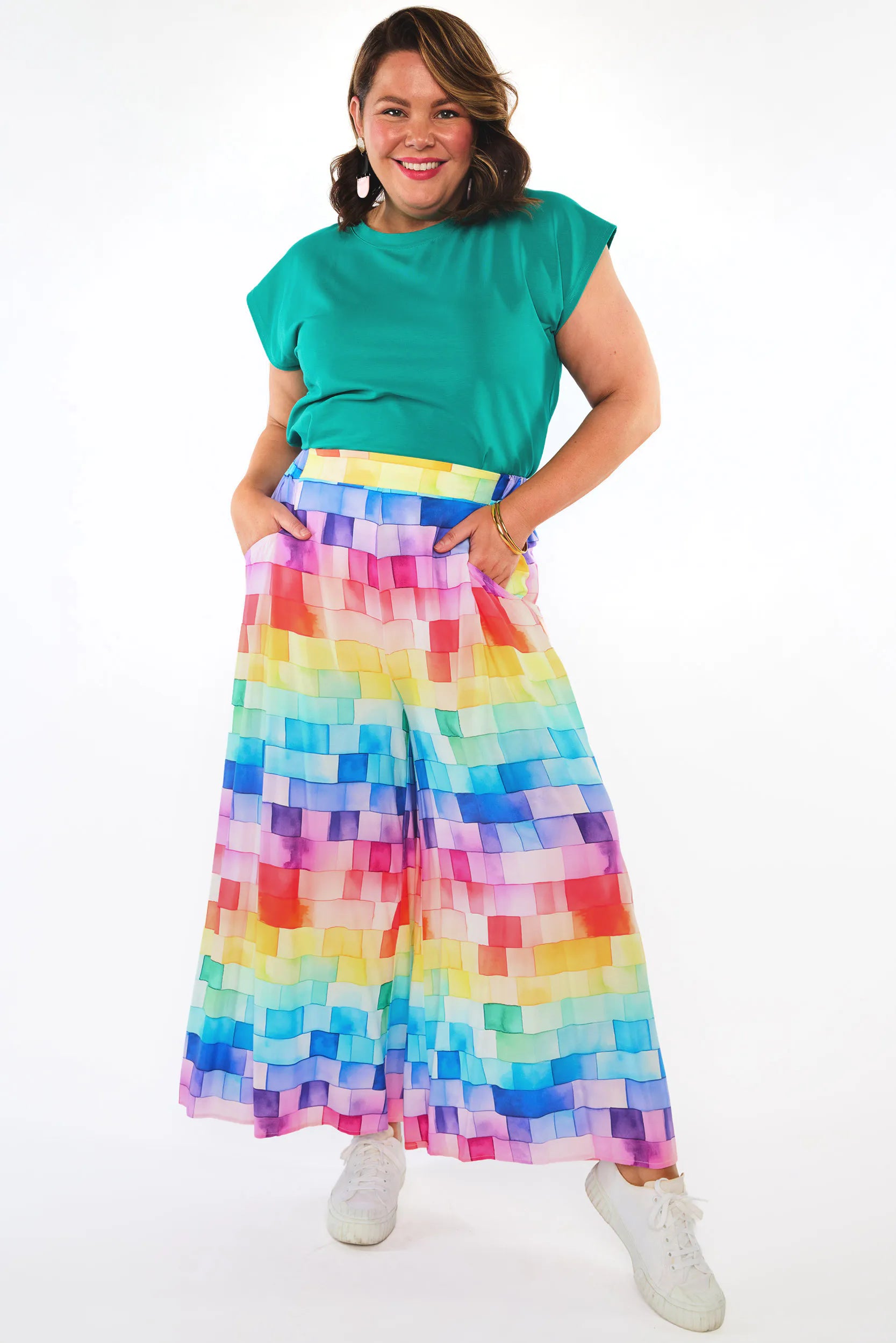 A woman modelling a vibrant, multicolour wide-leg pant with a pixelated block pattern, paired with a green top and white sneakers, against a plain white studio background.