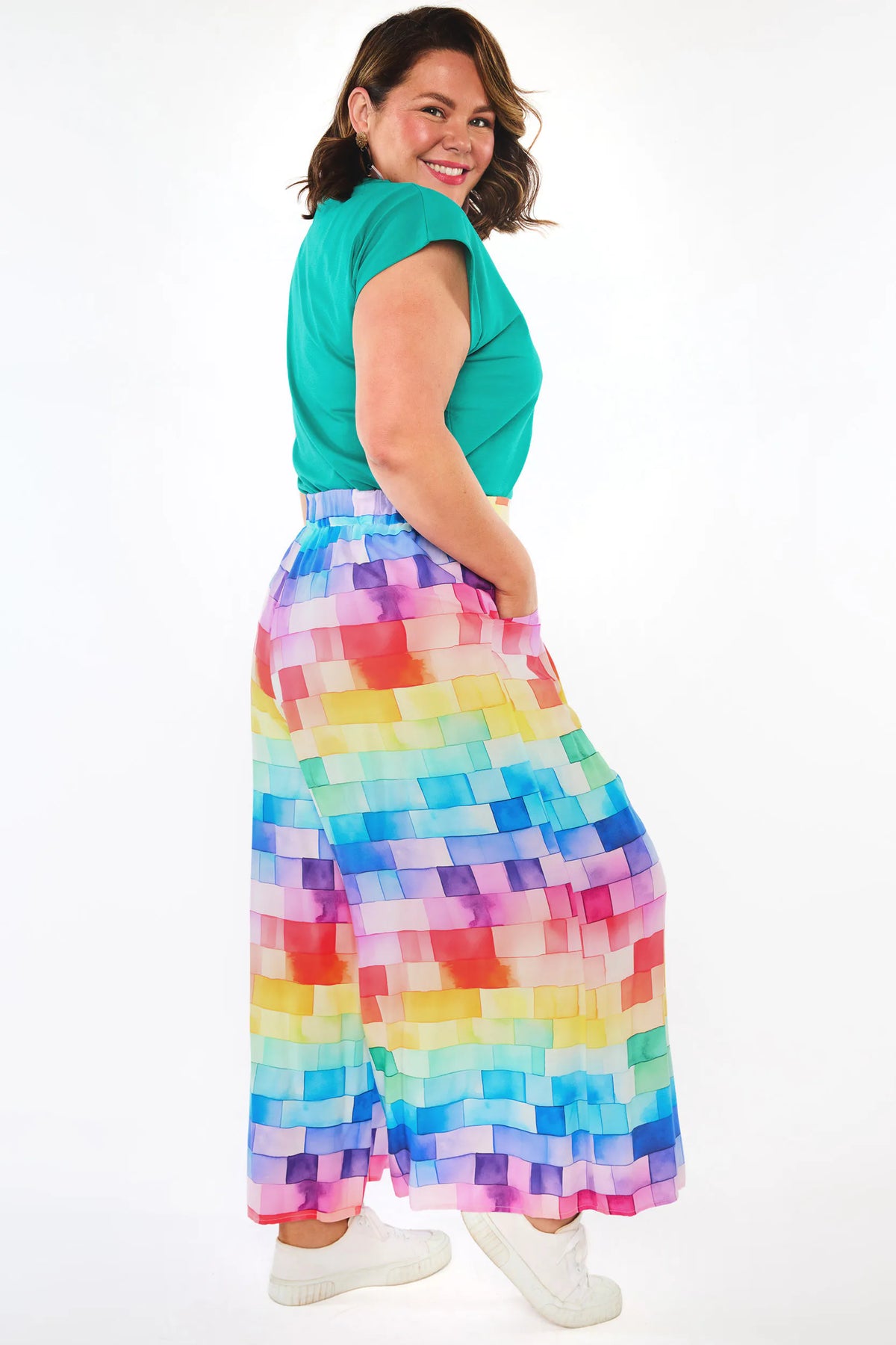 A woman modeling a vibrant, multicolor pixelated print maxi skirt paired with a green top and white sneakers, photographed against a solid white studio background.