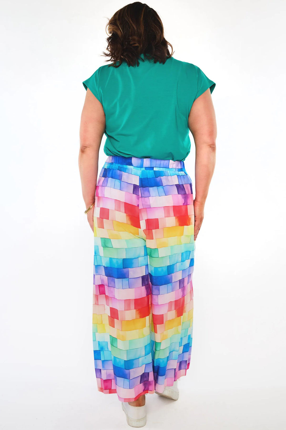 A woman viewed from behind wearing a bright green short-sleeve top and high-waisted, wide-leg pants with a colorful pixelated block pattern in shades of blue, pink, yellow, green, and purple, set against a plain white studio background.