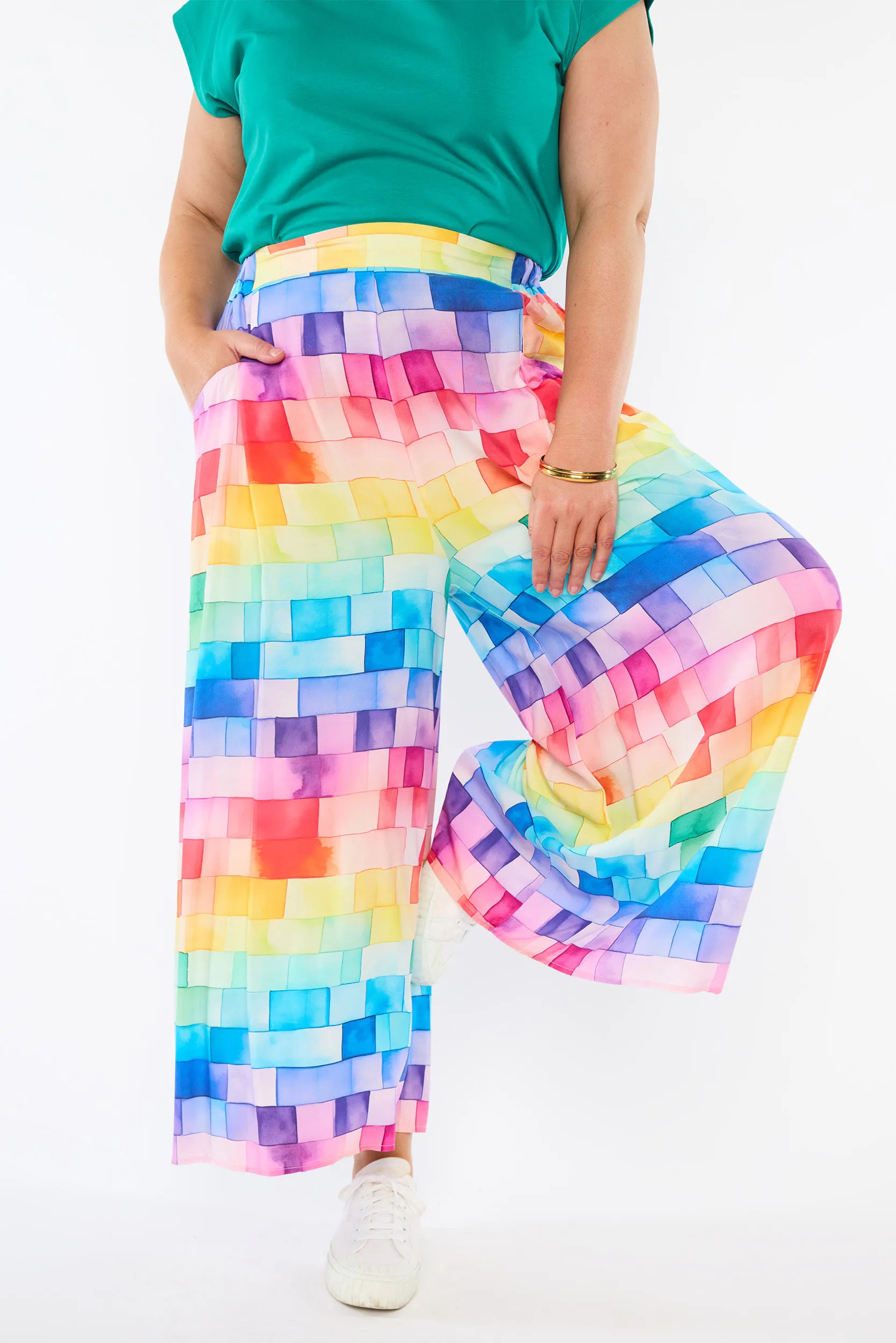 A person wearing wide-leg cropped pants with a colorful rainbow pixelated pattern, paired with a green top and white sneakers, shown against a plain white background.
