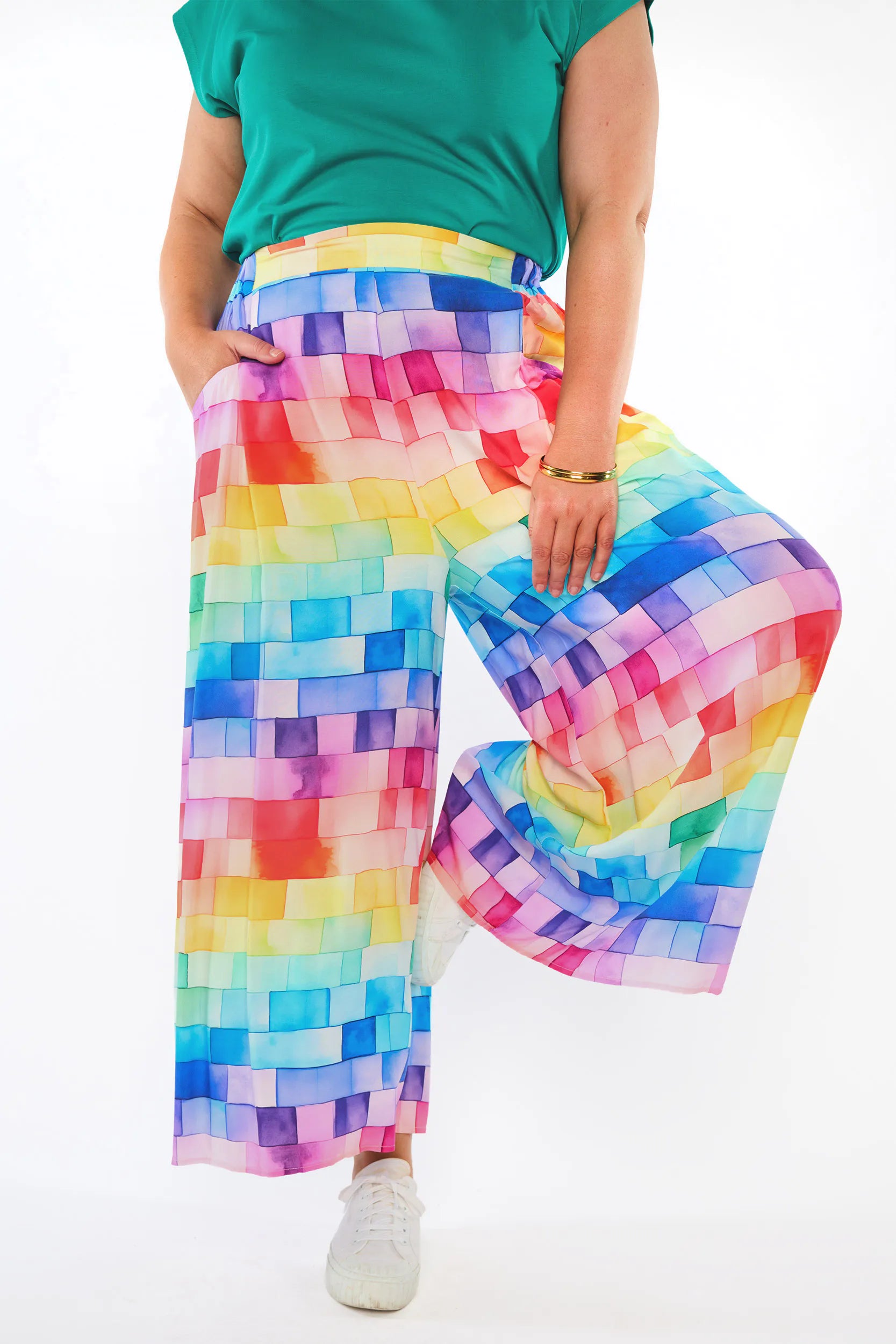 A person wearing wide-leg cropped pants with a colorful rainbow pixelated pattern, paired with a green top and white sneakers, shown against a plain white background.