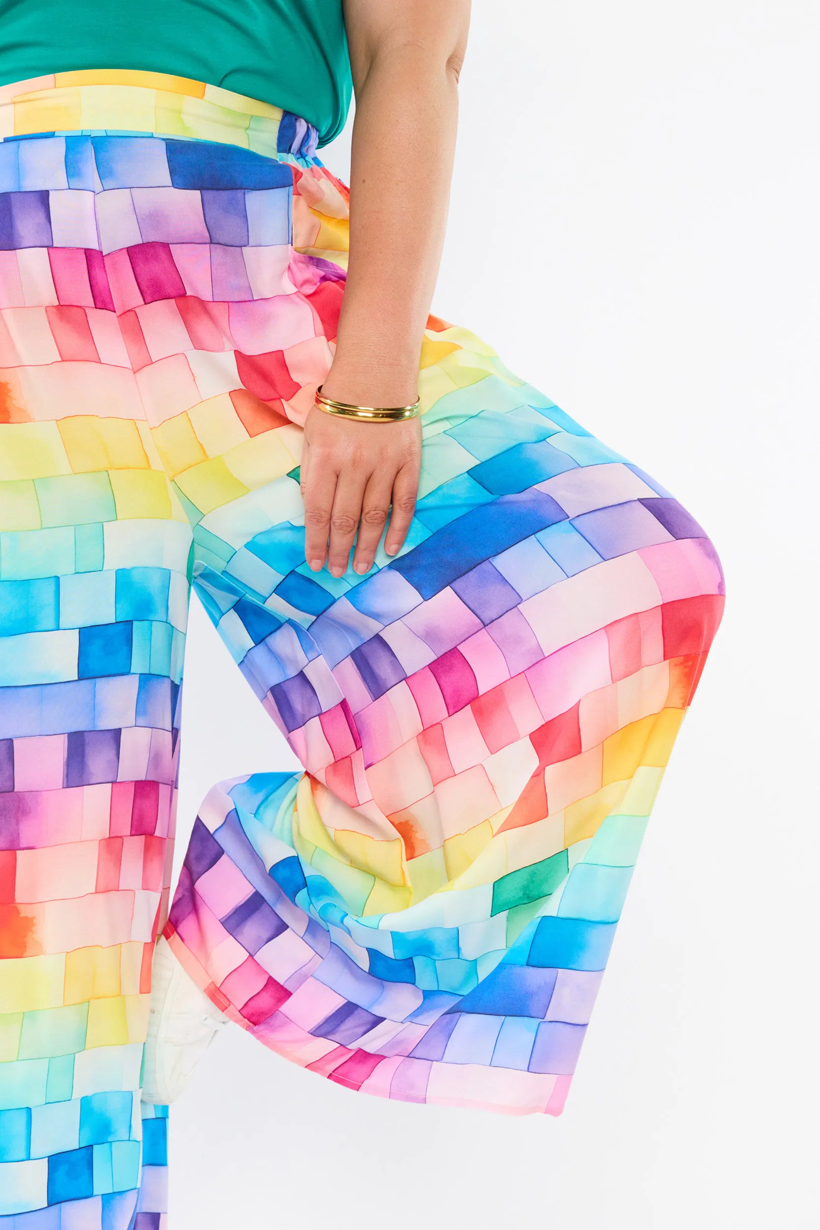 A close-up of a person wearing wide-leg pants with a colorful rainbow checkered pattern, paired with a green top. The person is shown from the waist down, with one leg bent, on a plain white background.