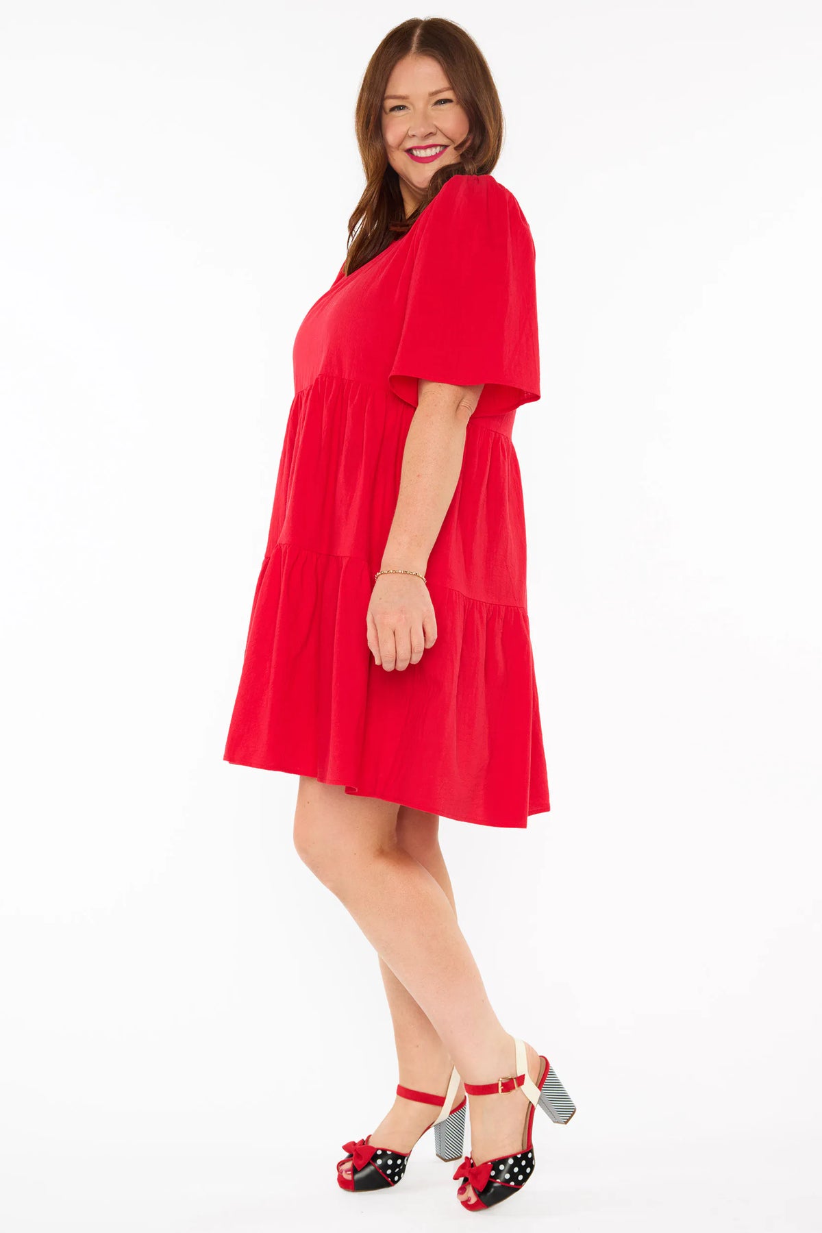 A woman modelling a bright red tiered mini dress with short puff sleeves and red bow heels against a plain white studio background.