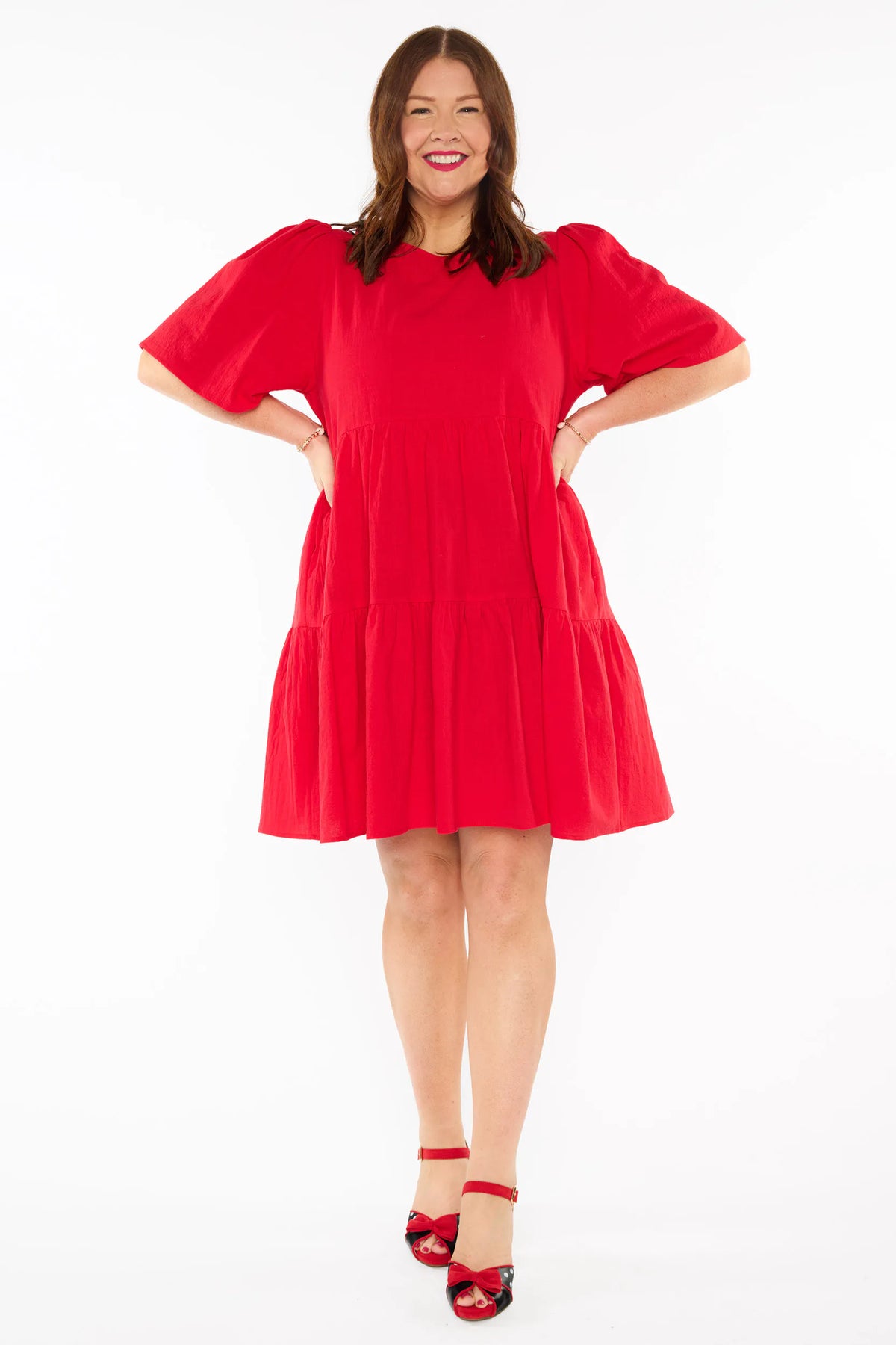 A woman modelling a bright red tiered mini dress with short puff sleeves and red bow heels against a plain white studio background.