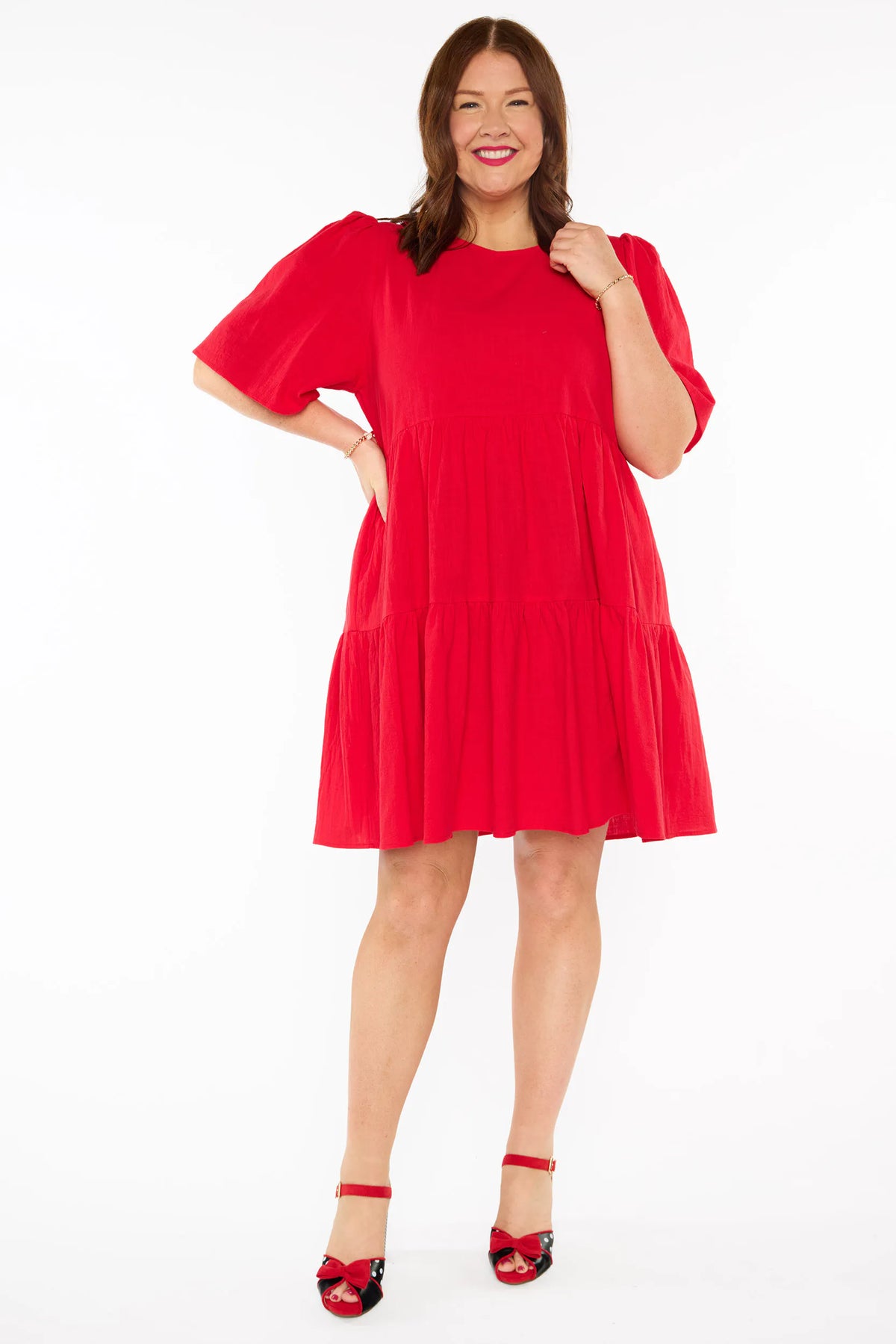 A woman modelling a bright red tiered mini dress with short puff sleeves and red bow heels against a plain white studio background.