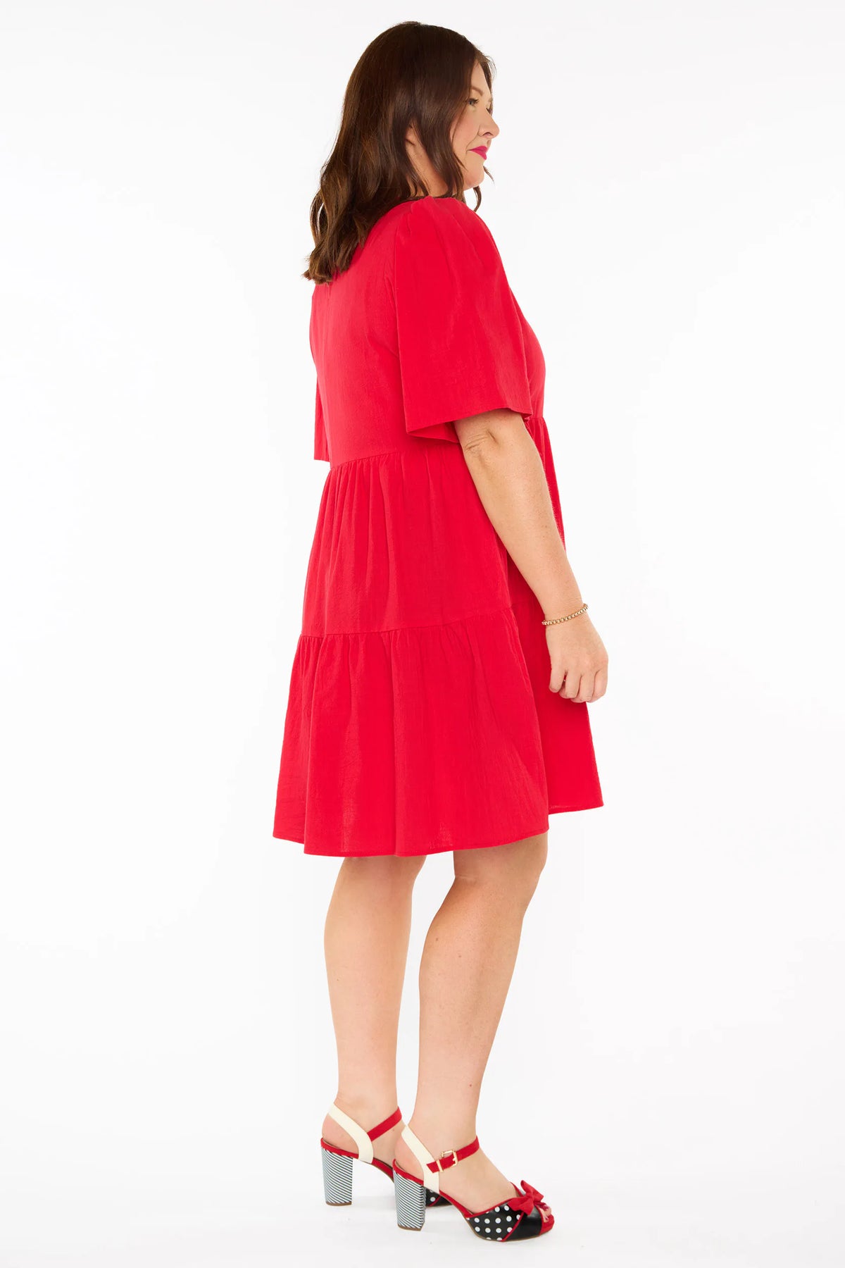 A woman modelling a bright red tiered mini dress with short puff sleeves and red bow heels against a plain white studio background.