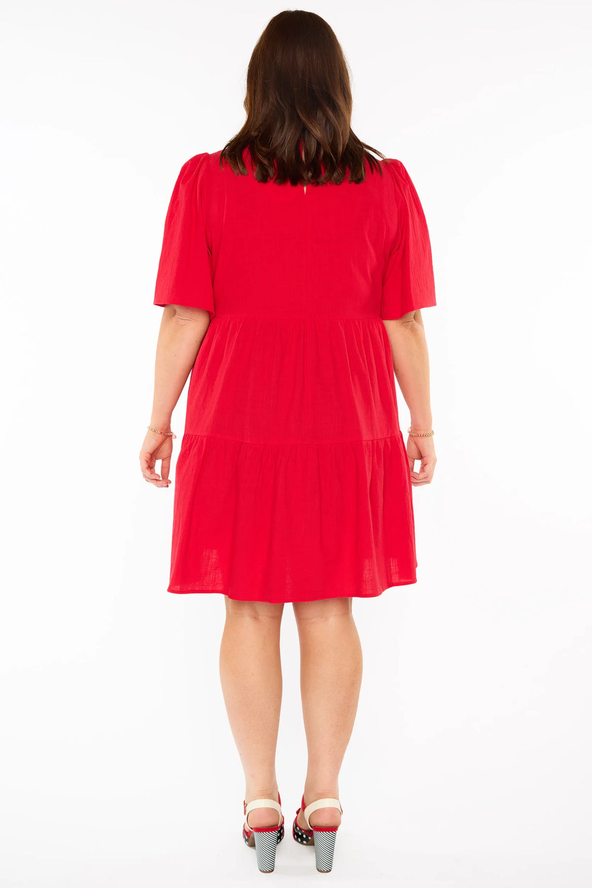 A back view of a woman modelling a bright red tiered mini dress with short puff sleeves and red bow heels against a plain white studio background.