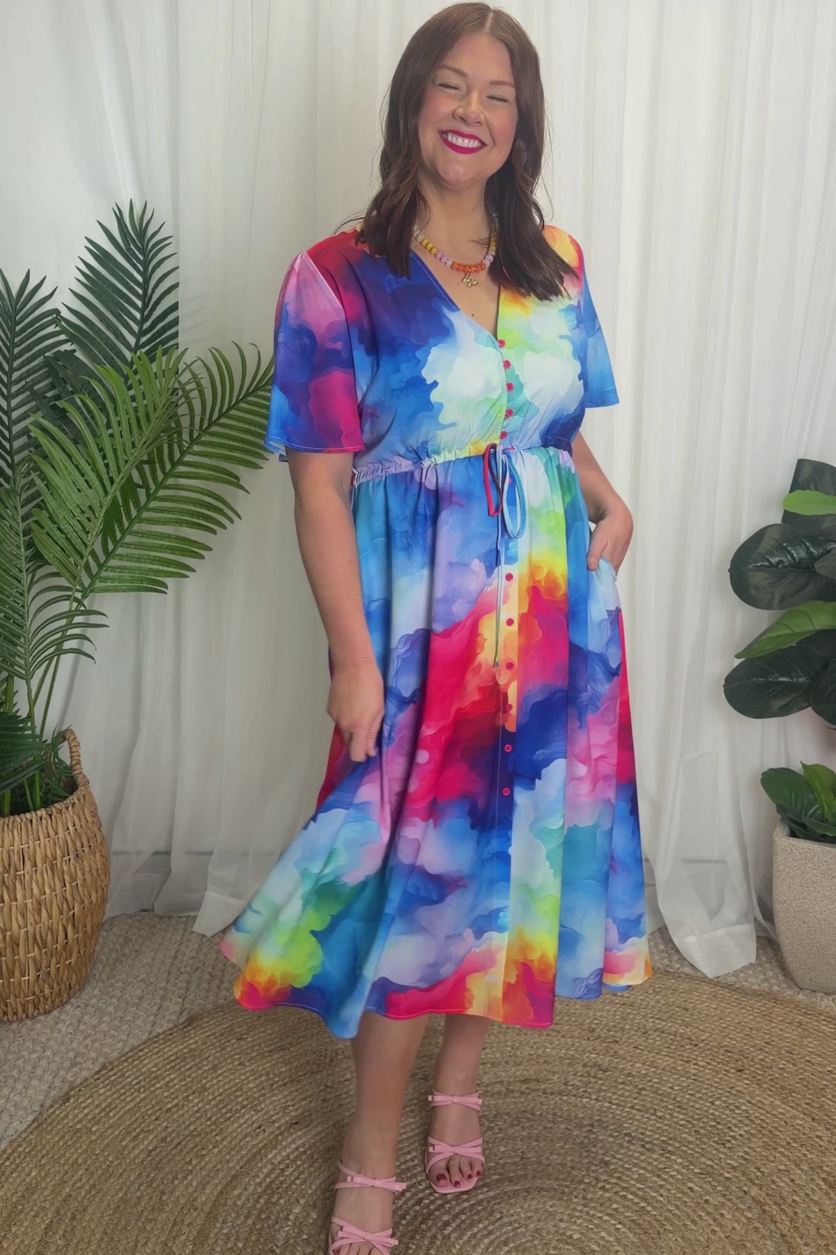 A woman modelling a short-sleeve midi dress with a vibrant watercolour tie-dye pattern in shades of blue, pink, yellow, green, and purple, set against a plain white studio background.