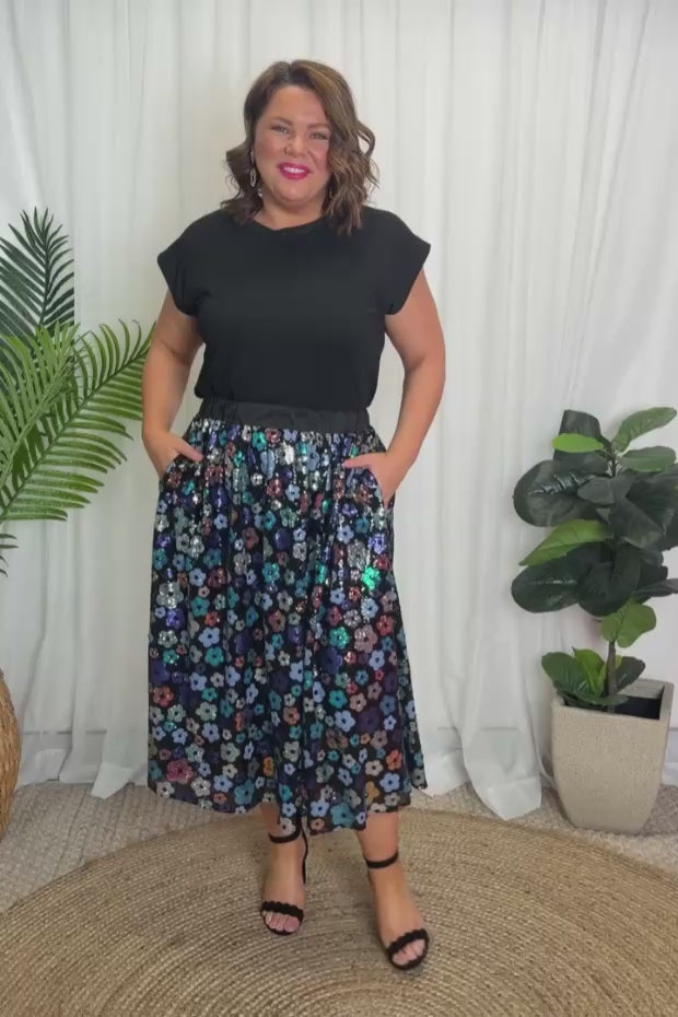 A woman modelling a short-sleeve black top, with a sequin knee length skirt featuring a mix of blue tonal coloured flowers on a black background. Paired with black heels, against a plain white studio background.