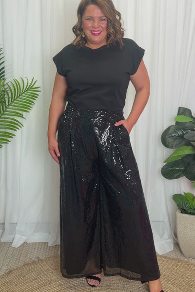 A woman modelling a black short sleeves top and a black sequin wide leg pant, standing against a plain white studio background with black heels.