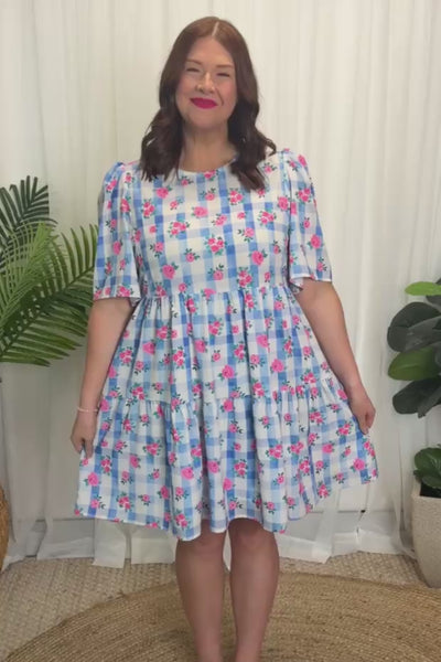 A woman modelling a short-sleeve, knee-length babydoll dress with a light blue and white gingham background covered in pink little roses, paired with pink sneakers, against a plain white curtain background.