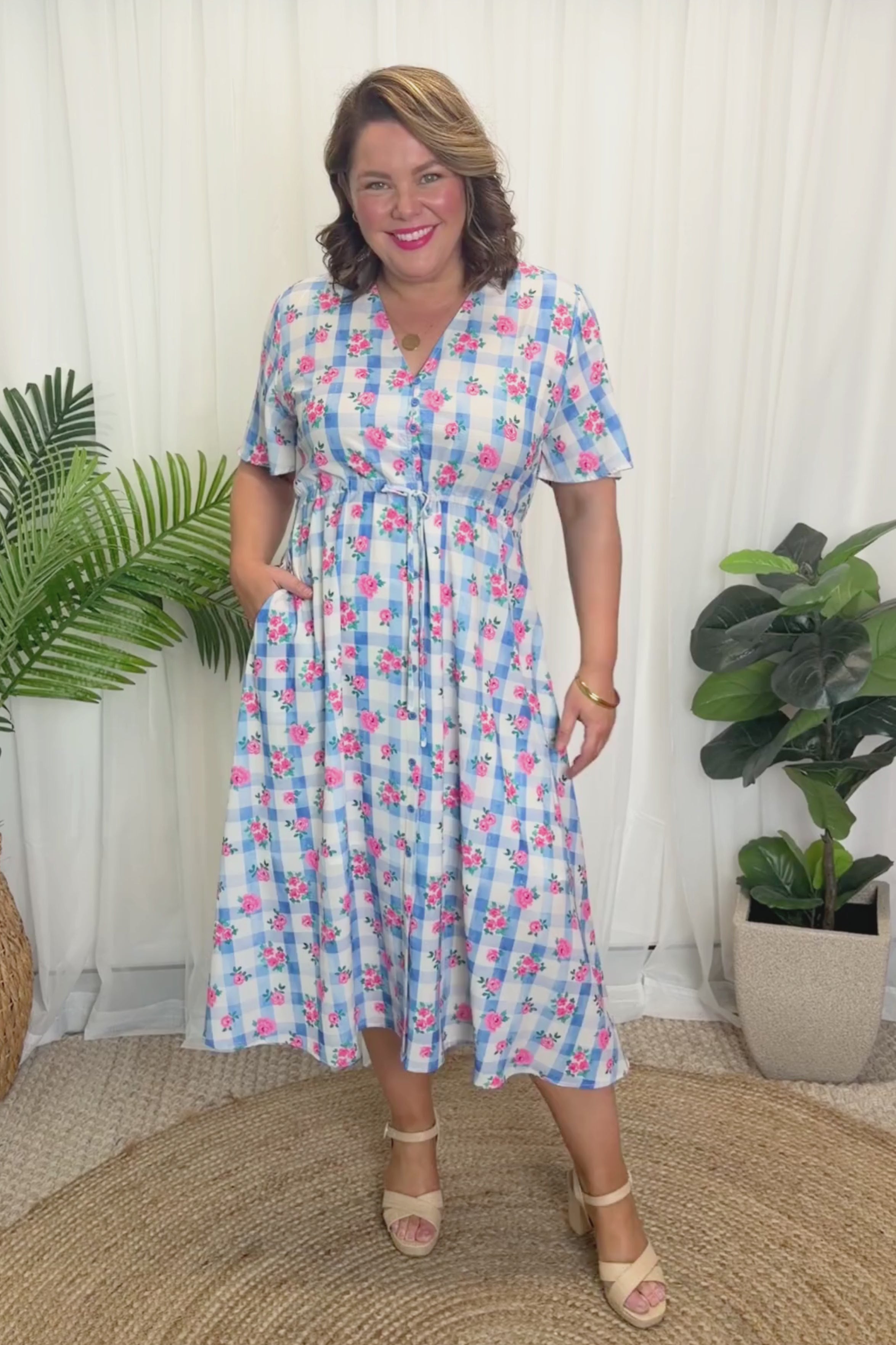 A woman modelling a short-sleeve, Midi-dress dress, with a button-up V-neckline & adjustable tie waist. A light blue and white gingham background covered in pink little roses, paired with cream heels, against a plain white curain background.