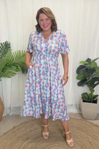 A woman modelling a short-sleeve, Midi-dress dress, with a button-up V-neckline & adjustable tie waist. A light blue and white gingham background covered in pink little roses, paired with cream heels, against a plain white curain background.