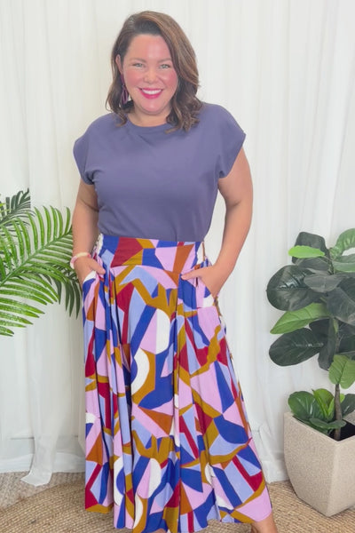 A woman modelling a sleeveless, high-waisted maxi skirt with a colourful geometric abstract print in blue, pink, red, and brown, paired with a solid navy blue top and beige platform sandals, against a plain white studio background.