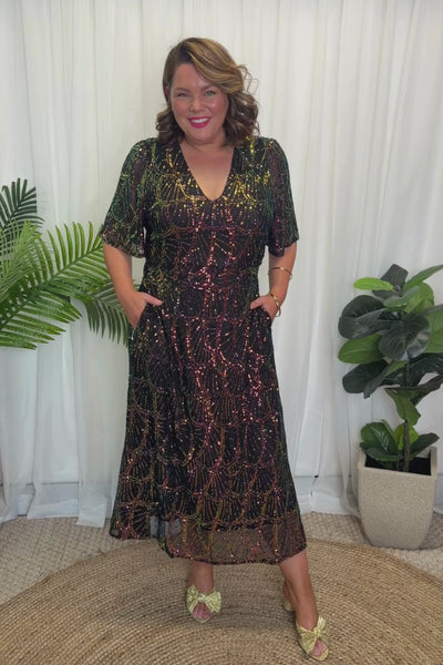 A woman modelling a short-sleeve V-neck midi dress covered in iridescent sequins that shift between green, pink, purple, and gold on a plain white background.