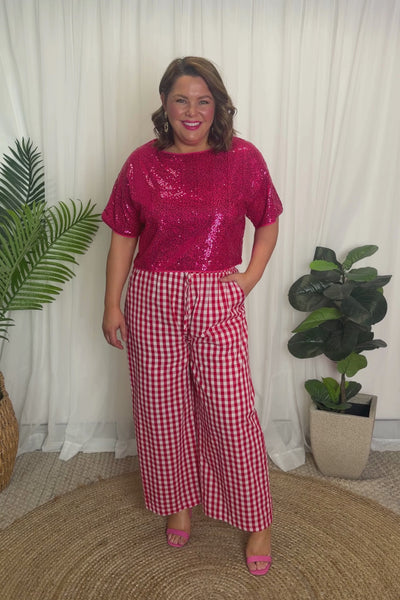 A woman modelling a two-piece outfit consisting of a bright pink sequins short-sleeve top and red-and-white gingham wide-leg pants, paired with pink heels, against a plain white background.